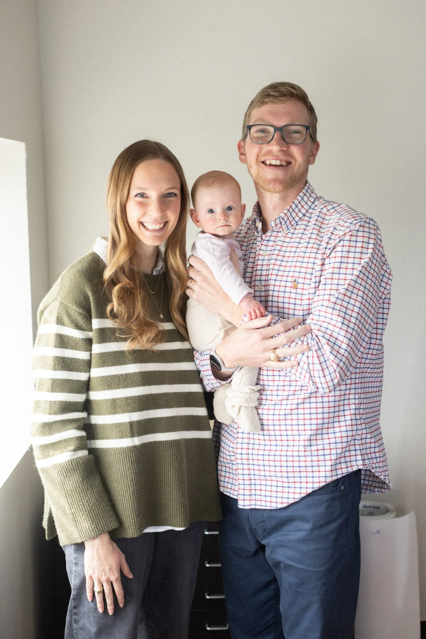 A smiling couple, a woman with long red hair wearing a green and white striped sweater and a man with glasses in a checkered shirt, hold a baby with light skin and no hair, dressed in a white onesie, inside a room with minimal decor.