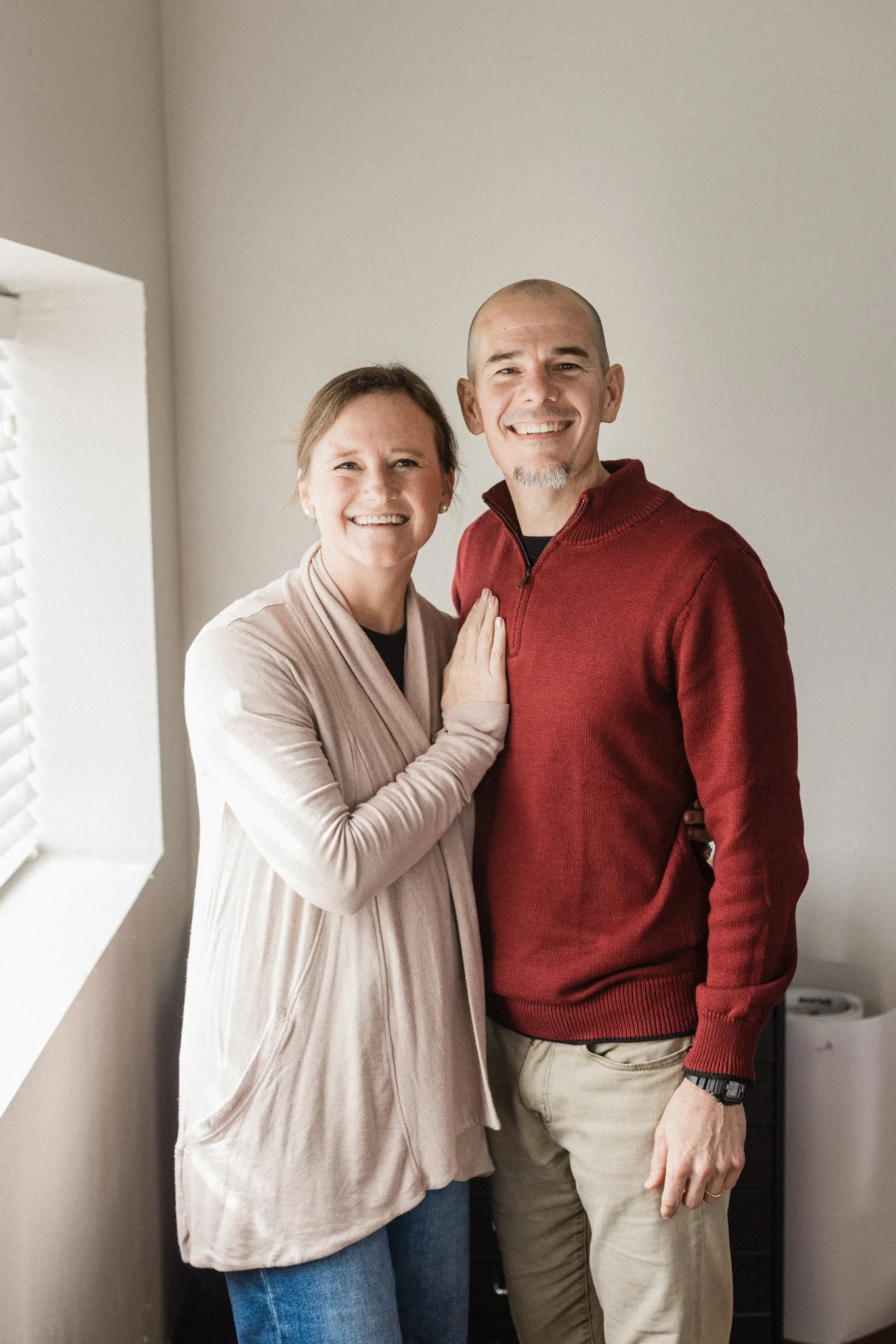 A woman and a man smiling with their arms around each other inside a room with plain walls and a partially visible window.