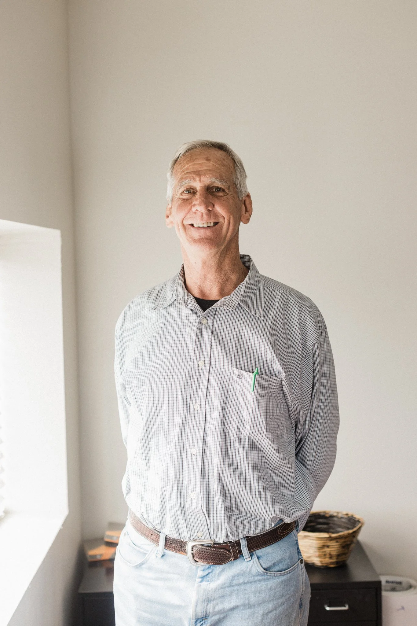 An elderly man with gray hair wearing a light checkered shirt, light jeans, and a brown belt, smiling and standing indoors near a black console table with a woven basket on it.