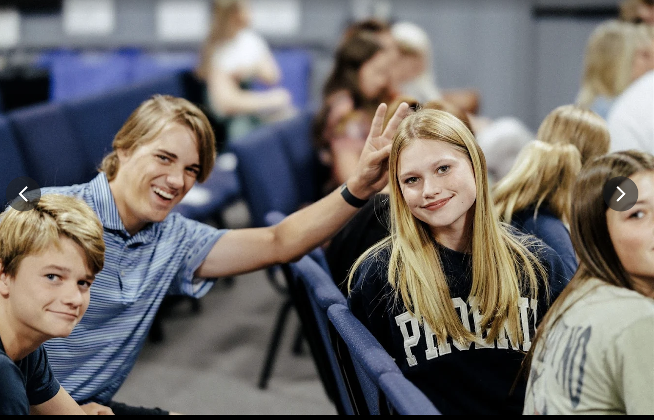 Charismatic young adults sitting in a lecture hall, engaging and smiling at the camera, with one person playfully touching another's ear.