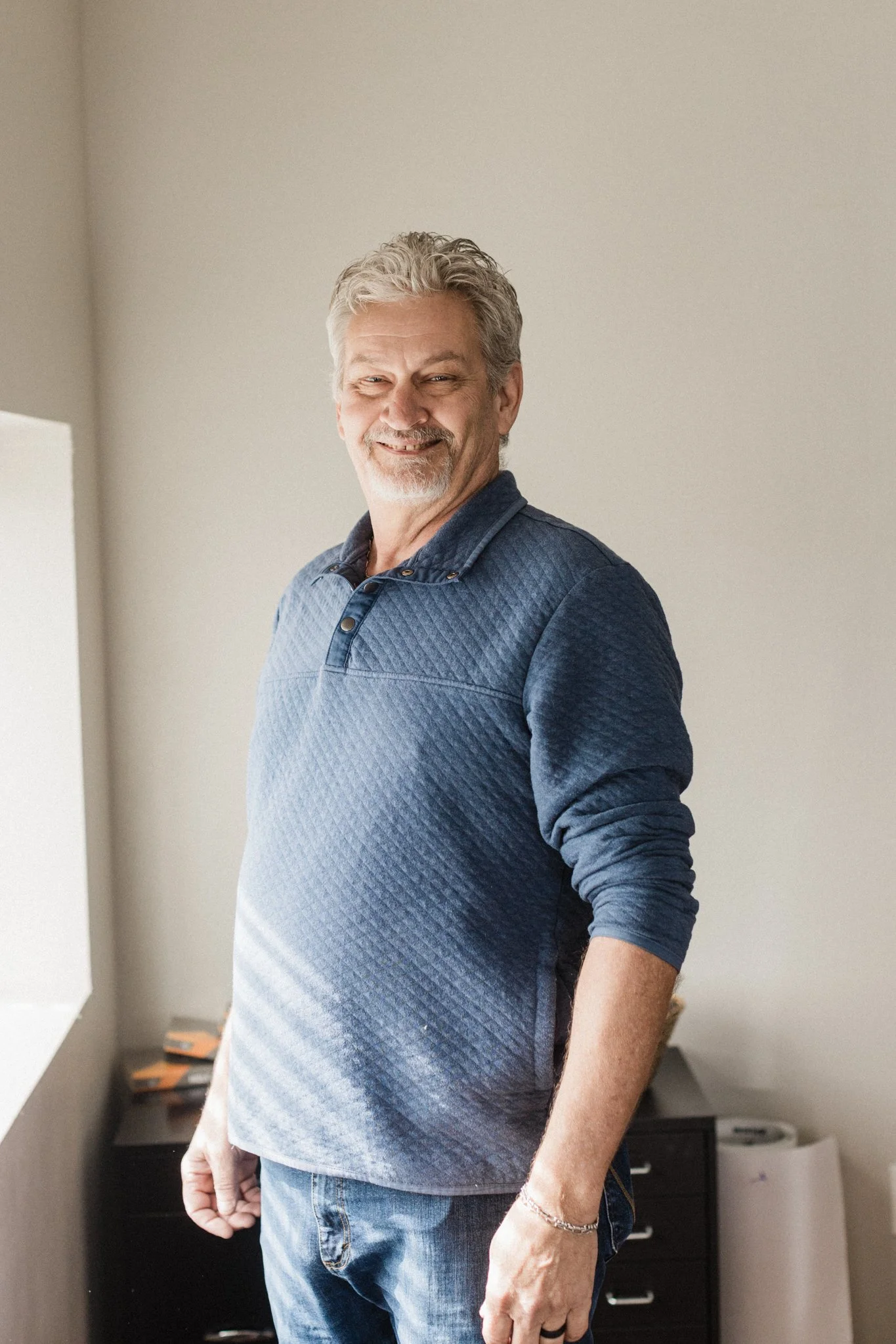 A smiling middle-aged man with gray hair and a beard wearing a blue textured long sleeve shirt, standing in a room with beige walls and a window.