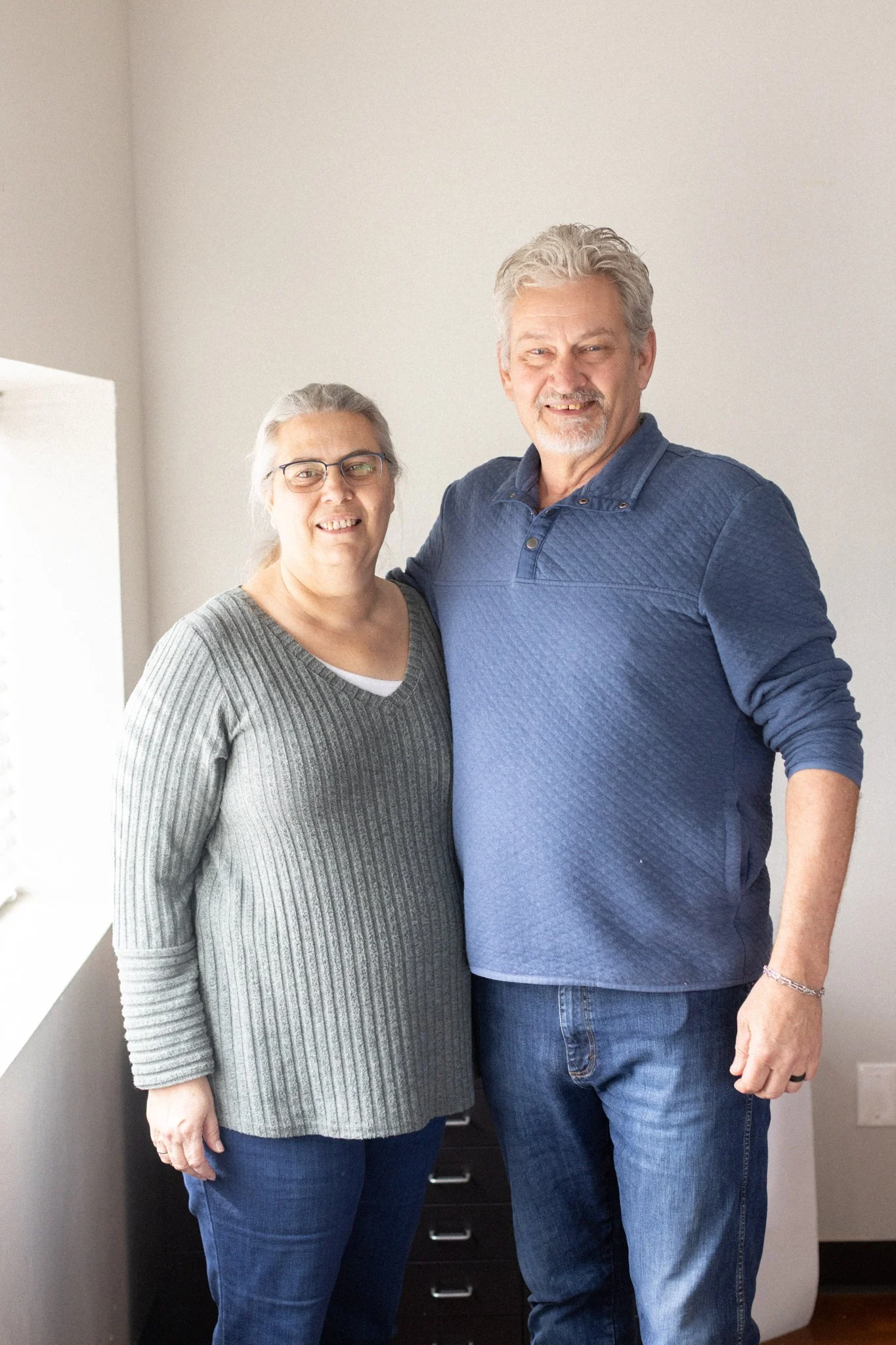 A woman and a man standing close together, smiling, in a room with plain white walls. The woman has gray hair, glasses, and is wearing a gray knitted sweater and jeans. The man has gray hair, a beard, and is wearing a blue long-sleeve button-up shirt and jeans.