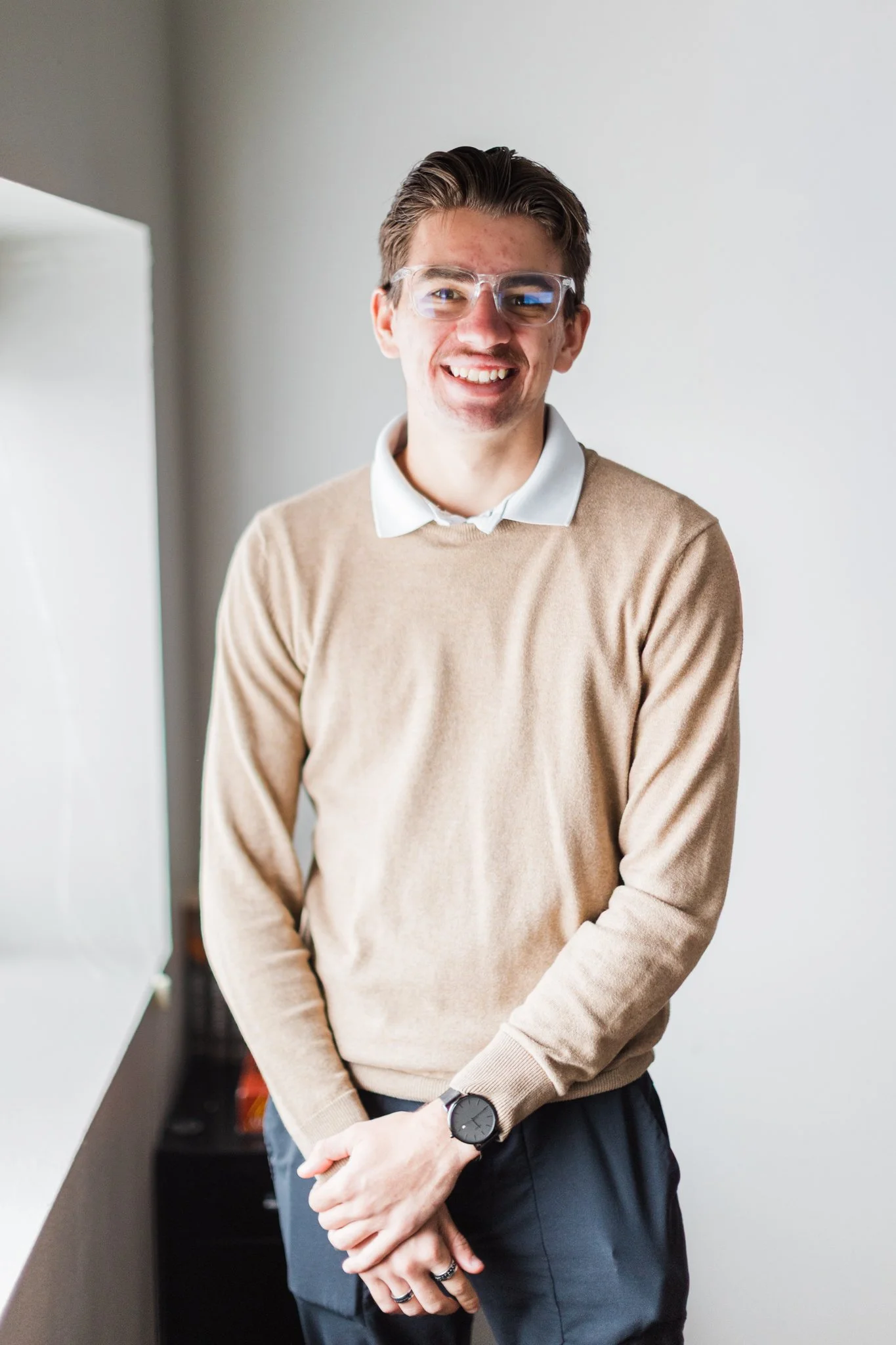 A young man with glasses smiling, wearing a beige sweater over a white collared shirt, with a watch on his left wrist, standing indoors near a white wall.