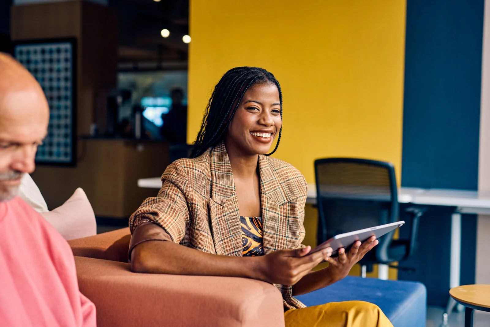 A woman sitting on a couch smiling and holding a tablet device in a modern office or cafe setting with a yellow wall and other people in the background.