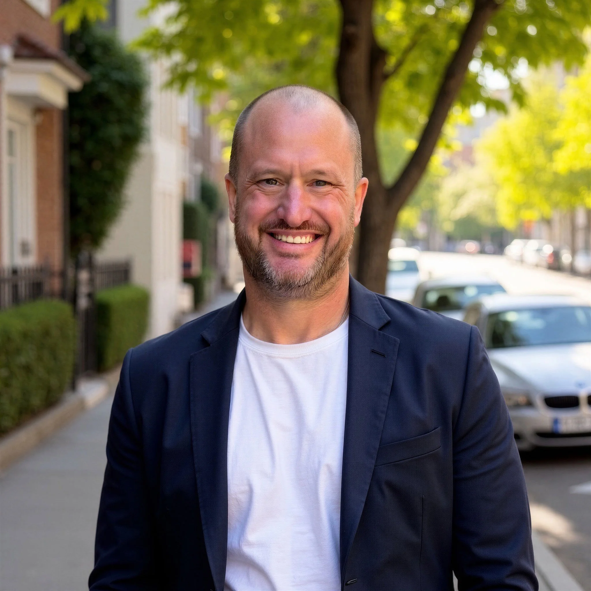 Portrait of a smiling man with a beard and a shaved head, wearing a dark blazer and white t-shirt, standing outdoors on a city street with trees and buildings in the background.