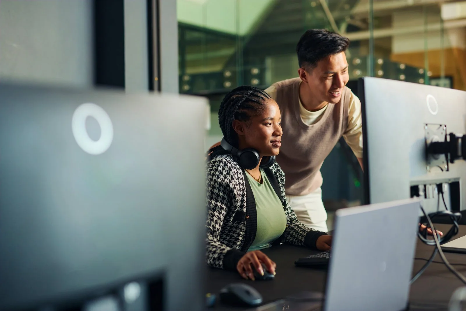 A woman with braided hair, wearing headphones around her neck, sitting at a computer with a man standing next to her, looking at the screen, in a modern office setting.