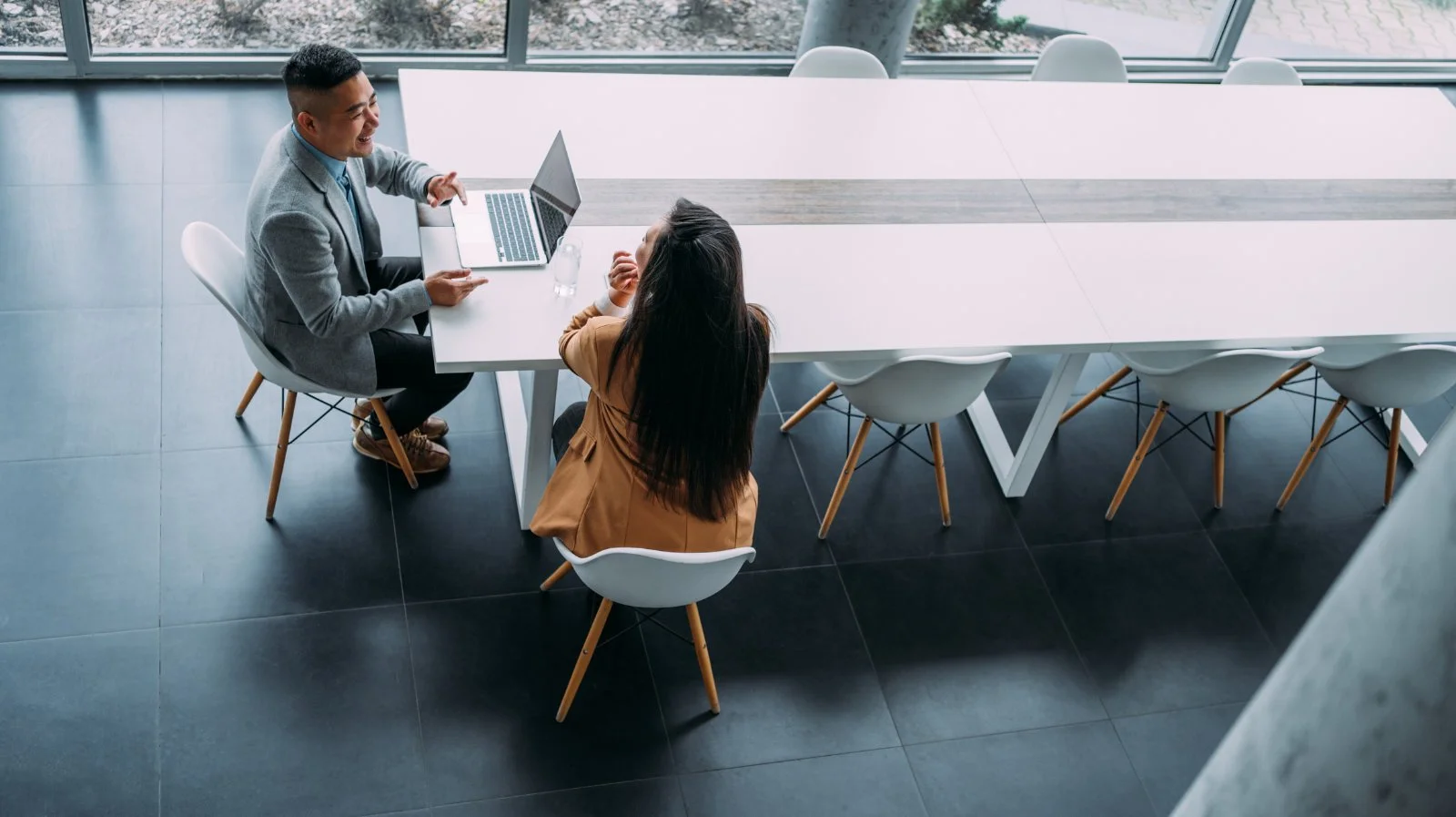 A man and woman sitting at a long white conference table in a modern office, talking and smiling. The man is wearing a gray blazer and the woman is wearing a tan blazer. There are chairs around the table and a water bottle in front of the woman.