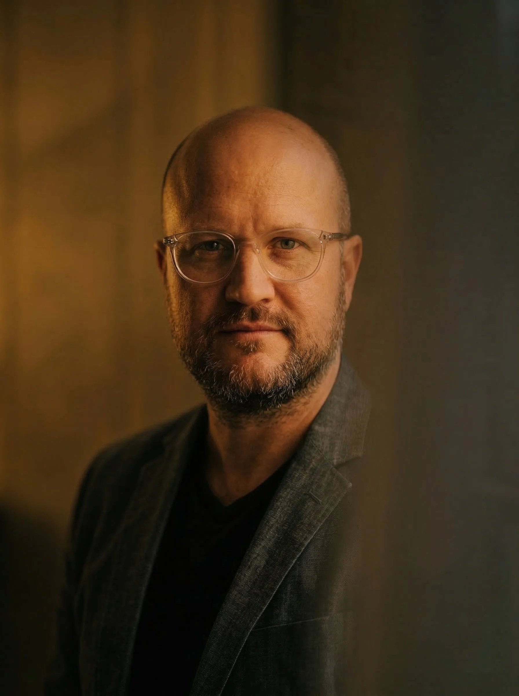 Close-up portrait of a middle-aged man with glasses, a beard, and a serious expression, dressed in a dark blazer, with a blurred background.