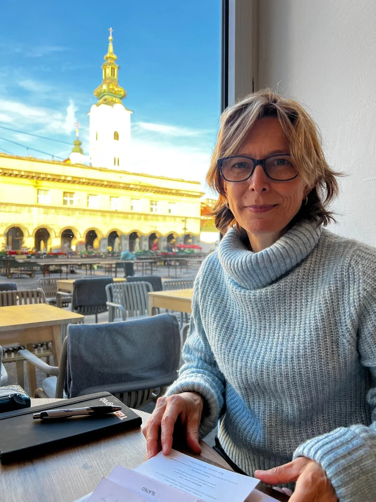Woman in a blue jumper with brown hair and glasses sits at a cafe table with a market square and historic building in the background.