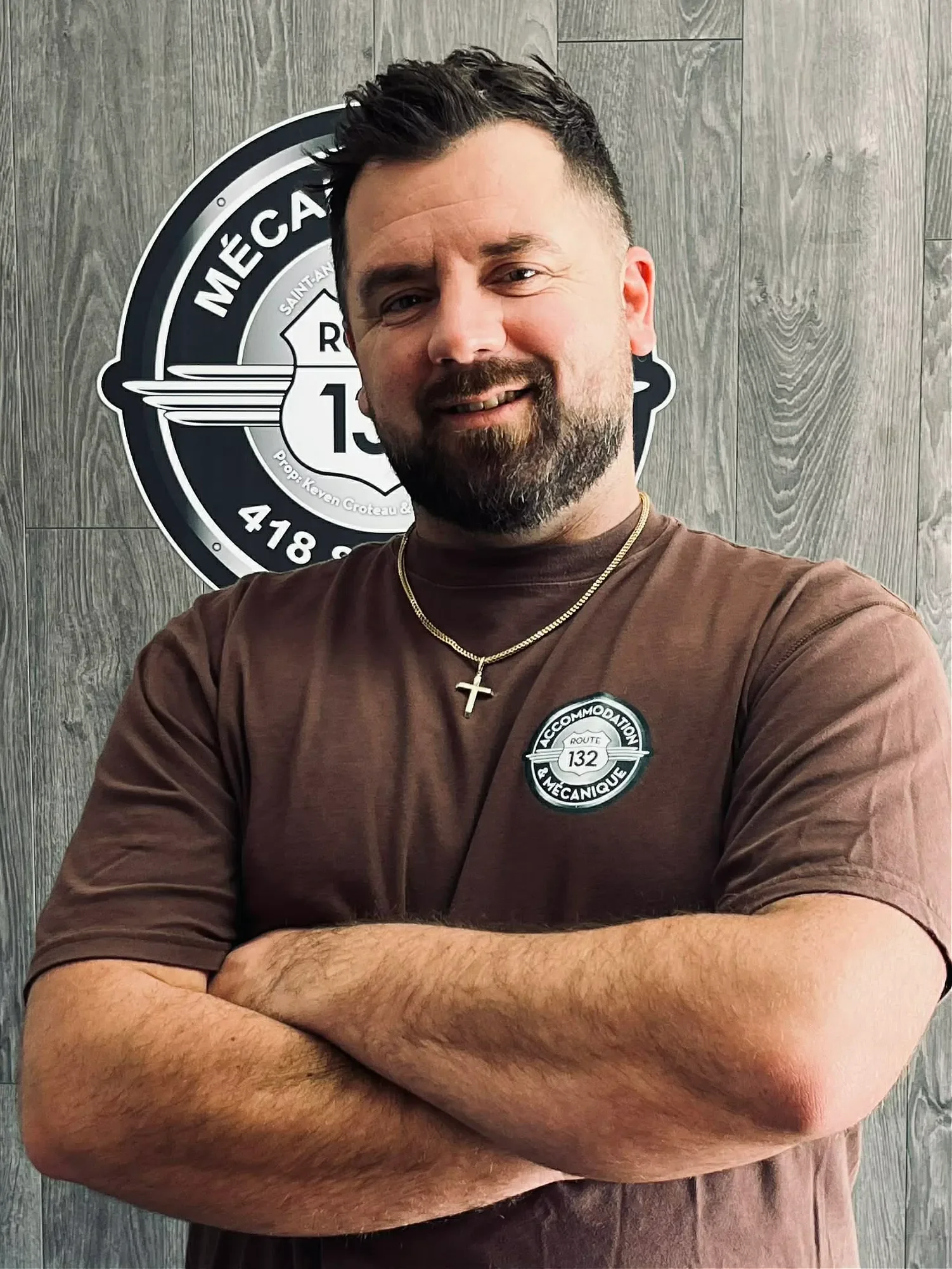 Un homme souriant avec une barbe, portant un t-shirt marron avec un badge de route 132 et une chaîne avec une croix, se tenant devant un mur en bois avec un logo noir et blanc de garage mécanique.