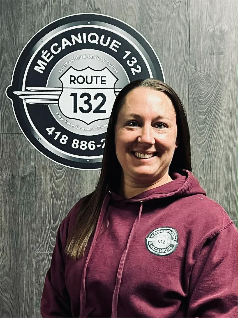 Femme souriante portant un sweat à capuche marron avec un badge de mécanique sur la poitrine, debout devant un panneau en bois avec un logo de route 132 et un numéro de téléphone.