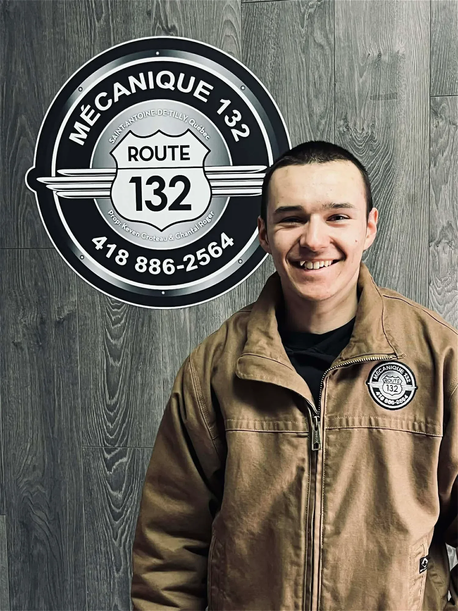 Un jeune homme souriant portant une veste marron avec un logo de route 132, debout devant un panneau en forme de cercle avec des informations sur la mécanique, Route 132, Québec, et un numéro de téléphone.