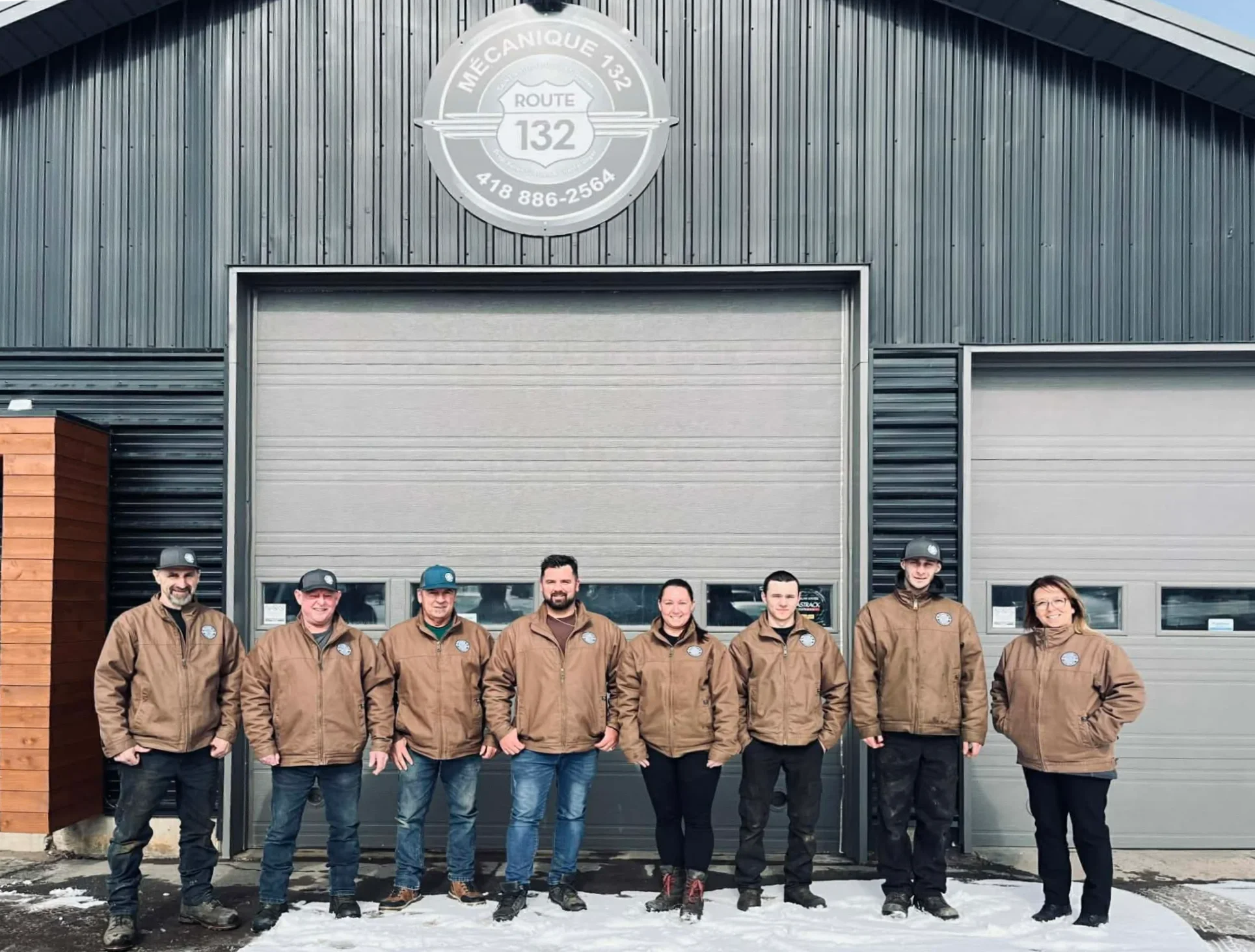 Groupe de neuf personnes debout devant un bâtiment en métal avec deux portes de garage, portant des vestes marron et des casquettes, par temps neigeux.