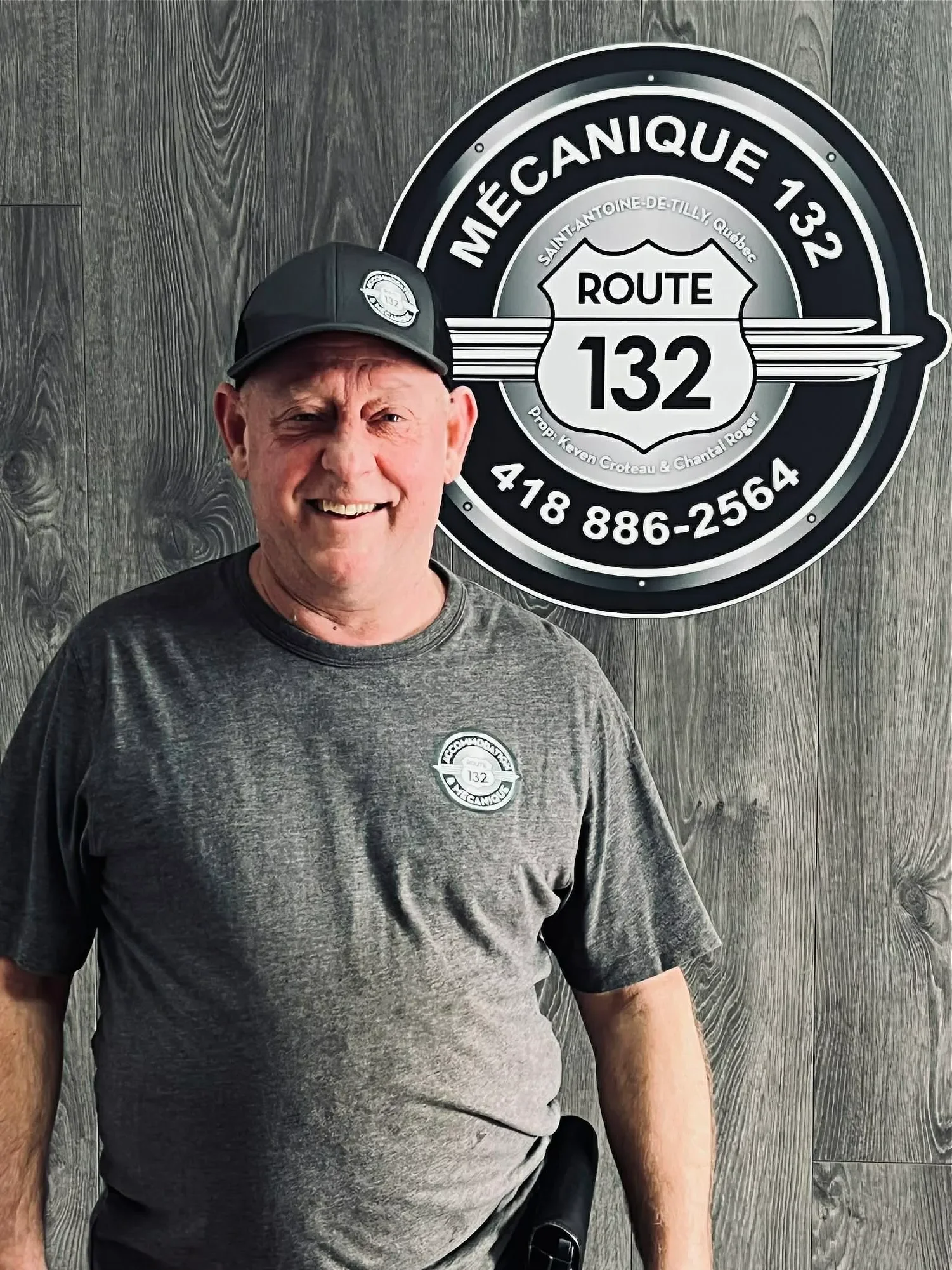 Un homme souriant portant un t-shirt gris et une casquette noire, se tenant devant un panneau de signalisation routière avec le texte "Méga Canique 132" et contact téléphonique, dans un décor en bois.