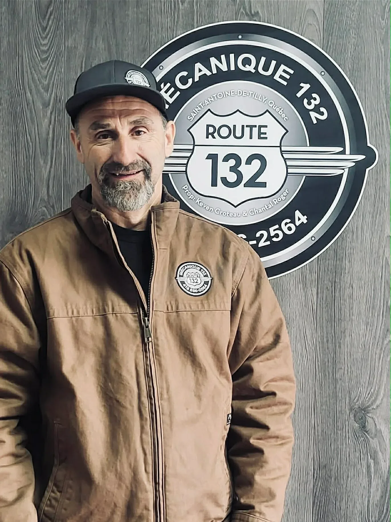Un homme avec une barbe grise portant une veste marron et une casquette noire, posant devant un panneau de route. Sur le panneau, il est écrit 'Route 132' et 'L'Équipement Canique 132'.