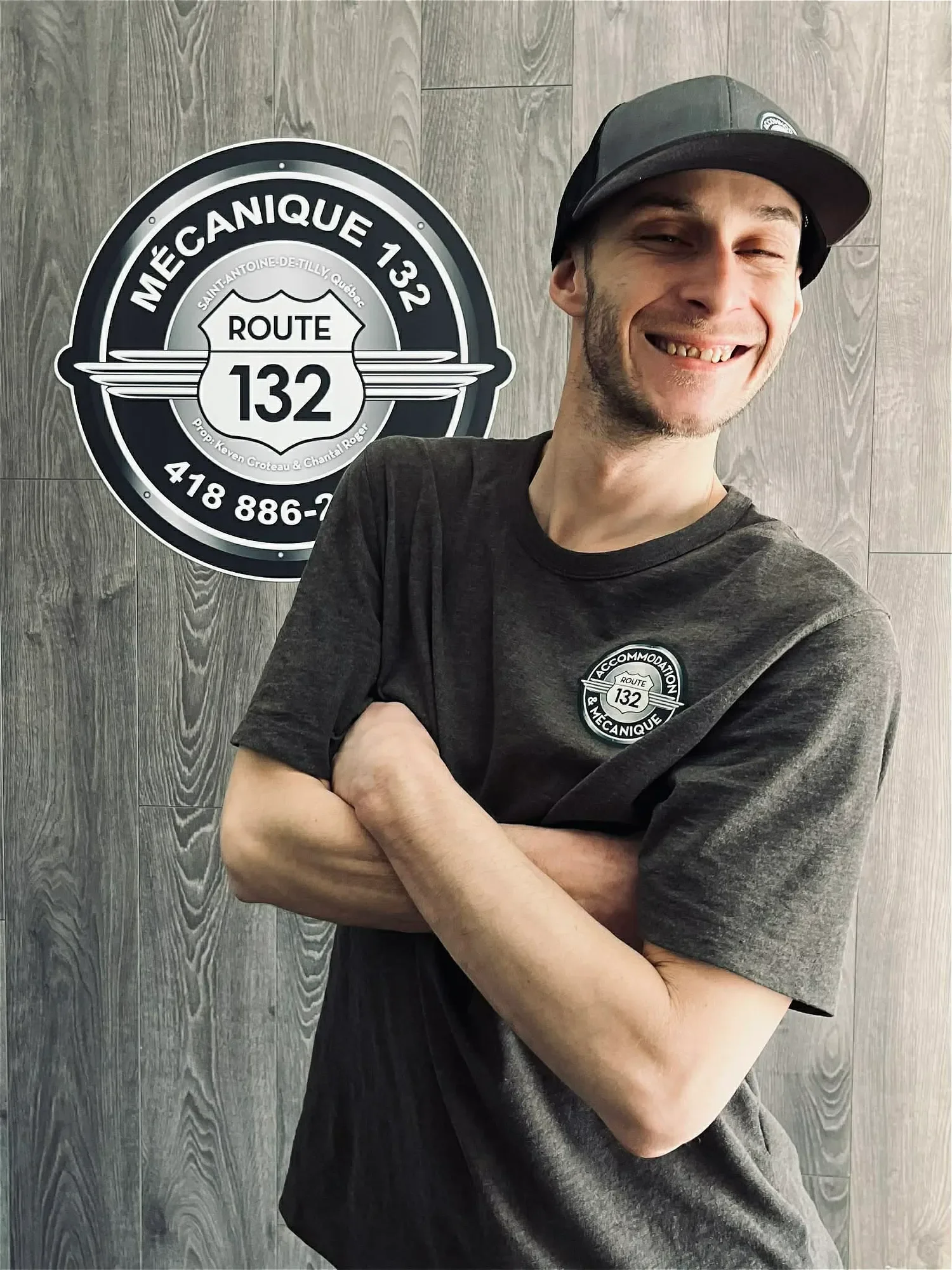 Un homme souriant portant un t-shirt et une casquette, avec un badge affichant 'Route 132 Mécanique' sur le mur en bois derrière lui.