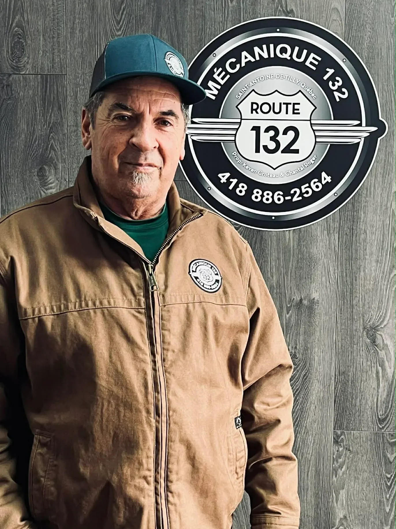 Homme portant une veste marron et une casquette bleue, se tenant devant un panneau avec le logo de la Route 132 et un fond en bois.
