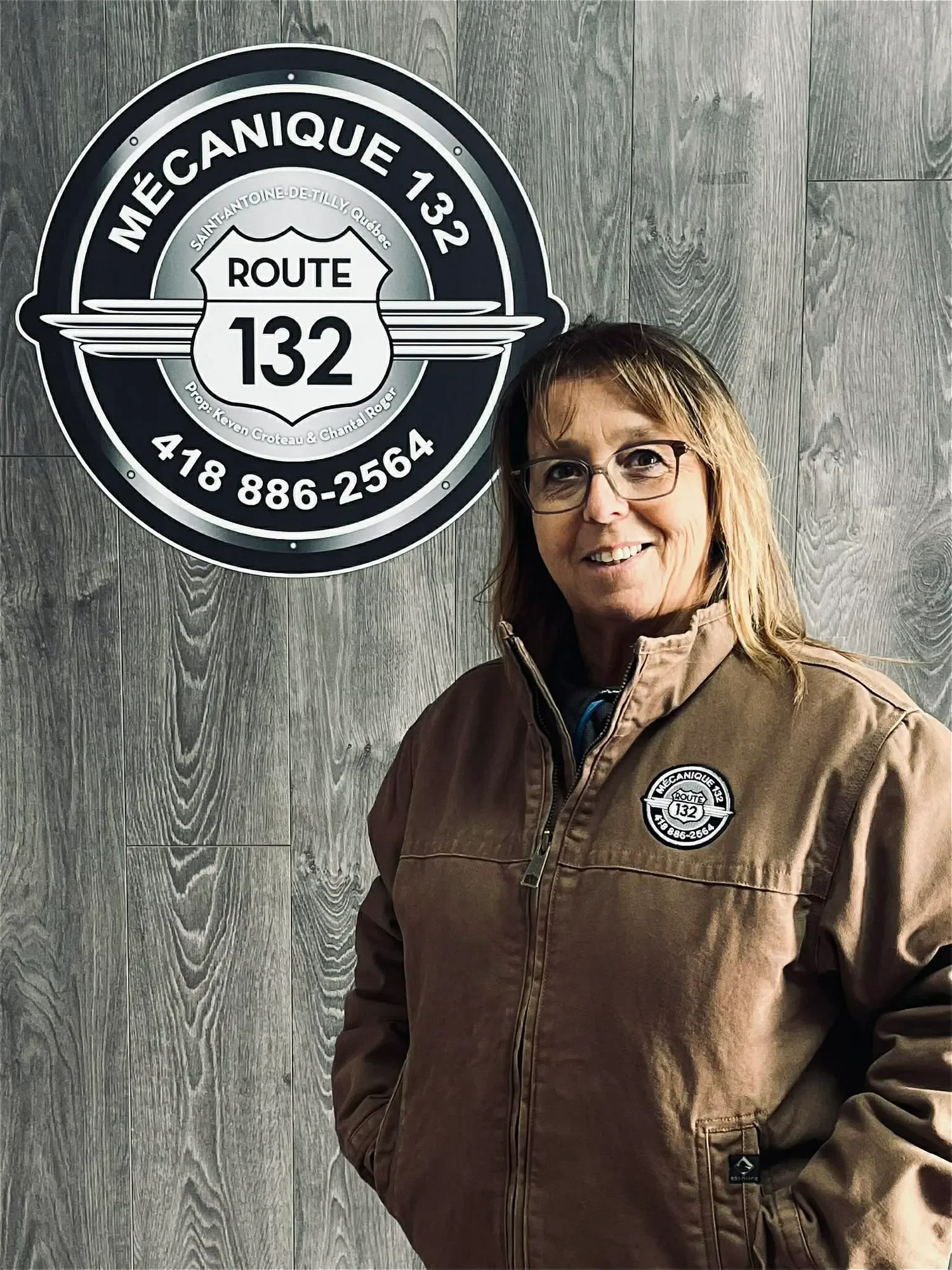 Une femme souriante porte une veste marron avec un logo de la route 132, devant un mur en bois avec une grande plaque d'immatriculation style badge de la route 132.