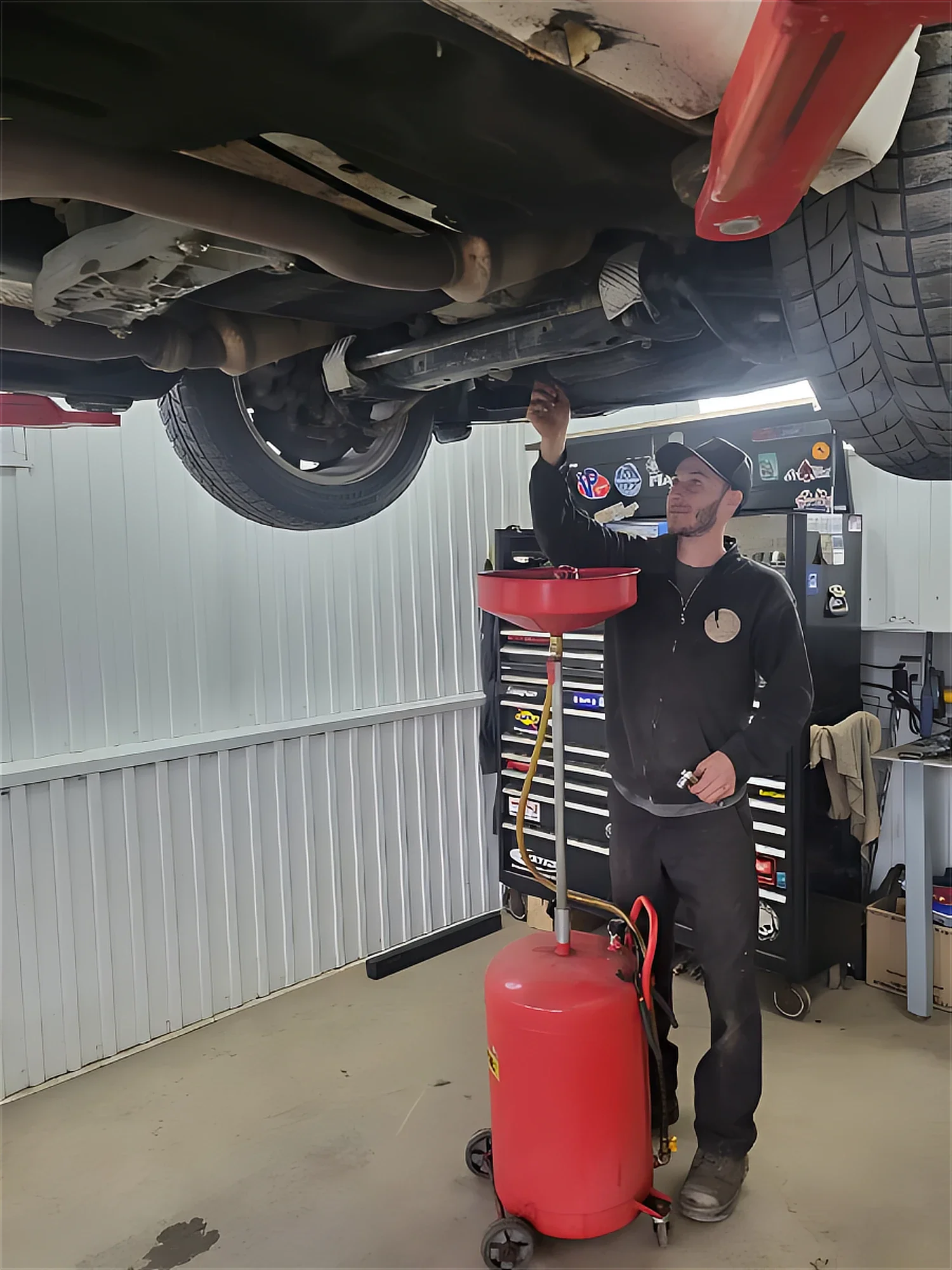 Un mécanicien effectue une inspection sous un véhicule levé dans un garage, utilisant un récipient rouge pour recueillir de l'huile ou un liquide.
