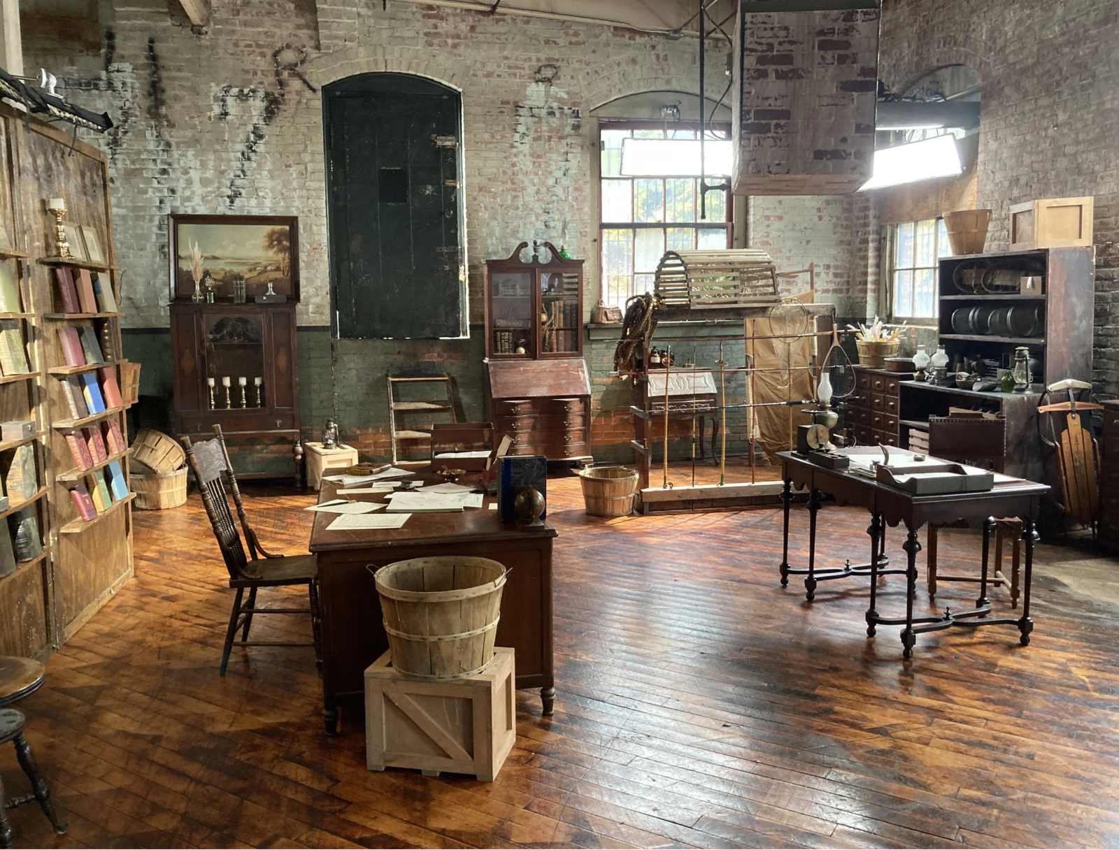 An antique office or study room with wooden furniture, bookshelves, desks, chairs, and various vintage objects. The room has brick walls, large windows, and hardwood floors.