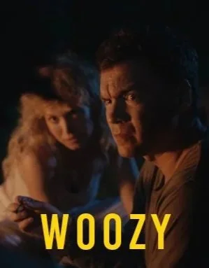 A man and a woman in a dark setting, with the man looking directly at the camera and the woman holding a phone. The word 'WOOZY' is written in yellow text at the bottom of the image.