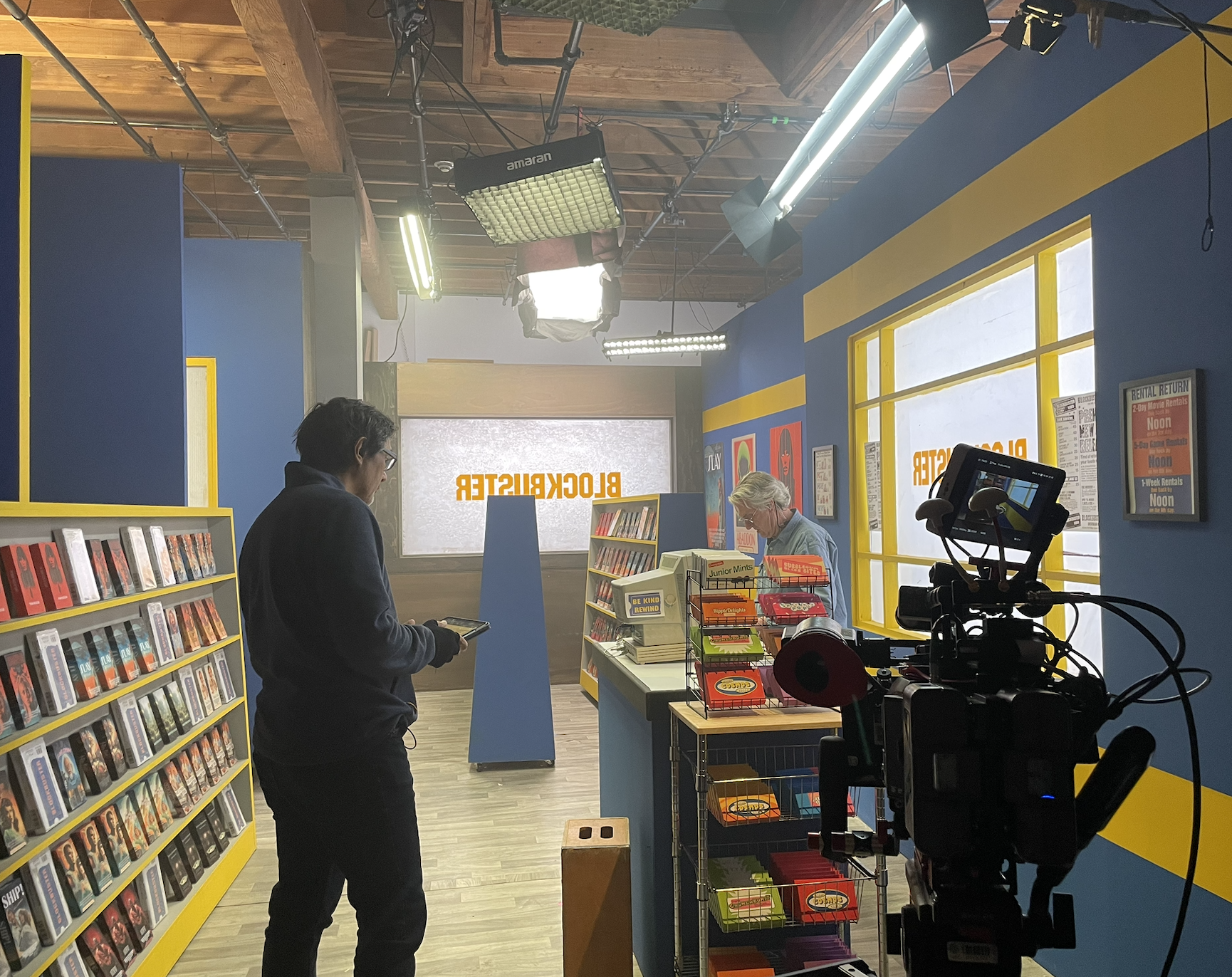 Interior of a bookstore with blue and yellow walls, featuring bookshelves filled with books, two people browsing, and filming equipment set up for recording.
