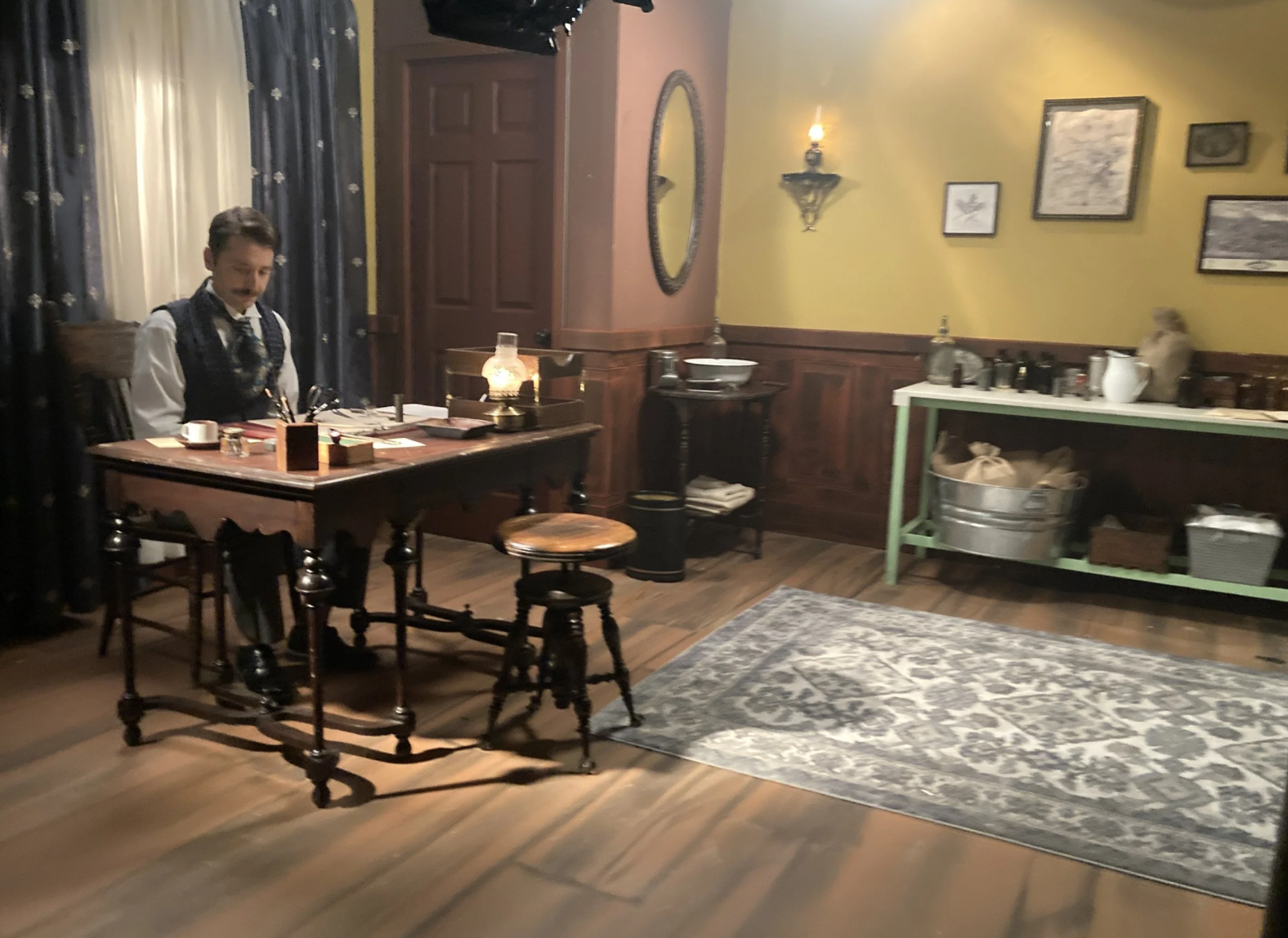 A person dressed in early 20th-century attire sitting at a wooden desk in a vintage-style room with framed pictures on yellow walls, a mirror, and a side table with supplies.