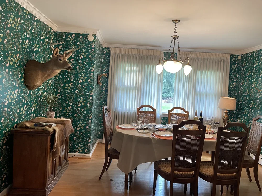 A dining room with a round table set for a meal, four chairs around it, green floral wallpaper, a chandelier, and a window with white curtains. There is a deer head mounted on the wall and a wooden sideboard with decorative items.