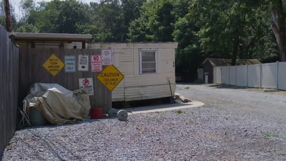 A small white mobile home surrounded by gravel, with multiple warning and parking signs attached to a wooden fence nearby, including warnings for children at play and no parking.