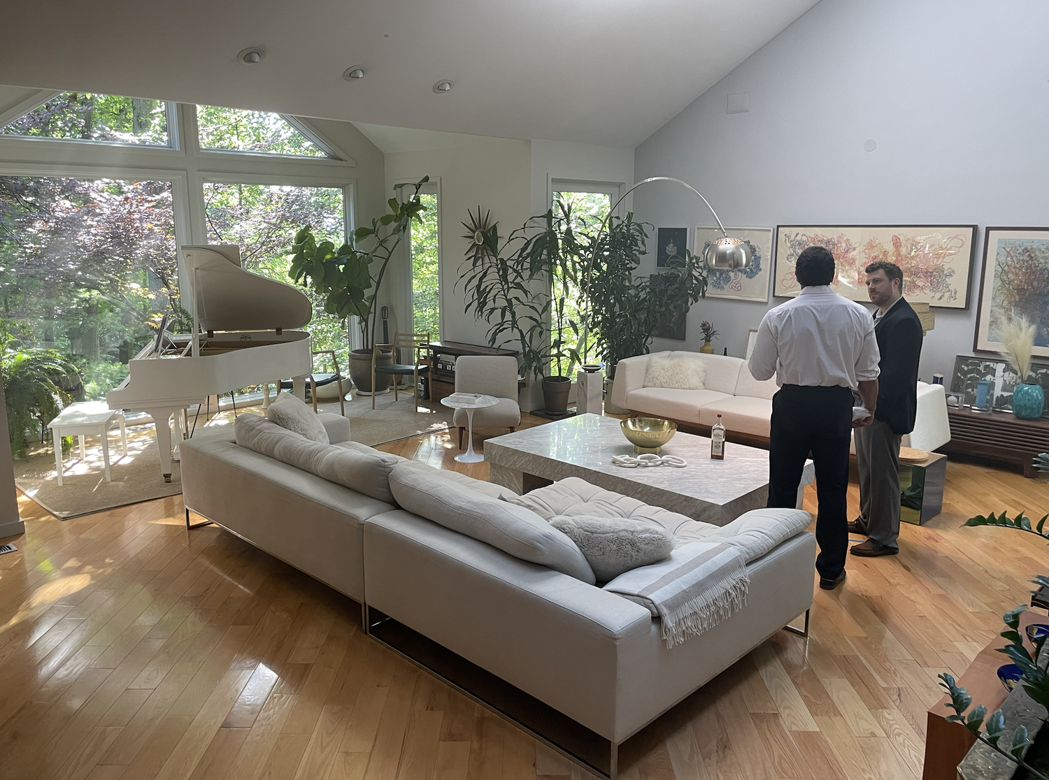 Living room with large windows, white couches, a grand piano, potted plants, and two men in conversation.