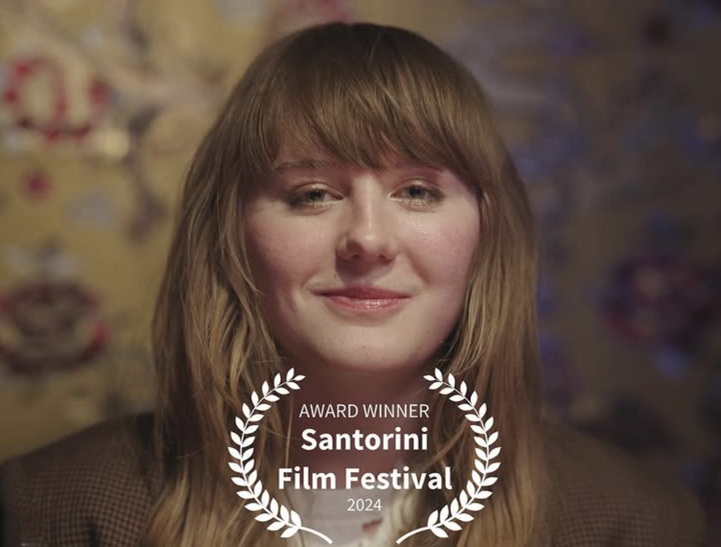 A woman with long, wavy, reddish-brown hair smiling gently, wearing a brown checkered blazer, with a blurred background and a laurel wreath graphic overlay stating "Award Winner Santorini Film Festival 2024".
