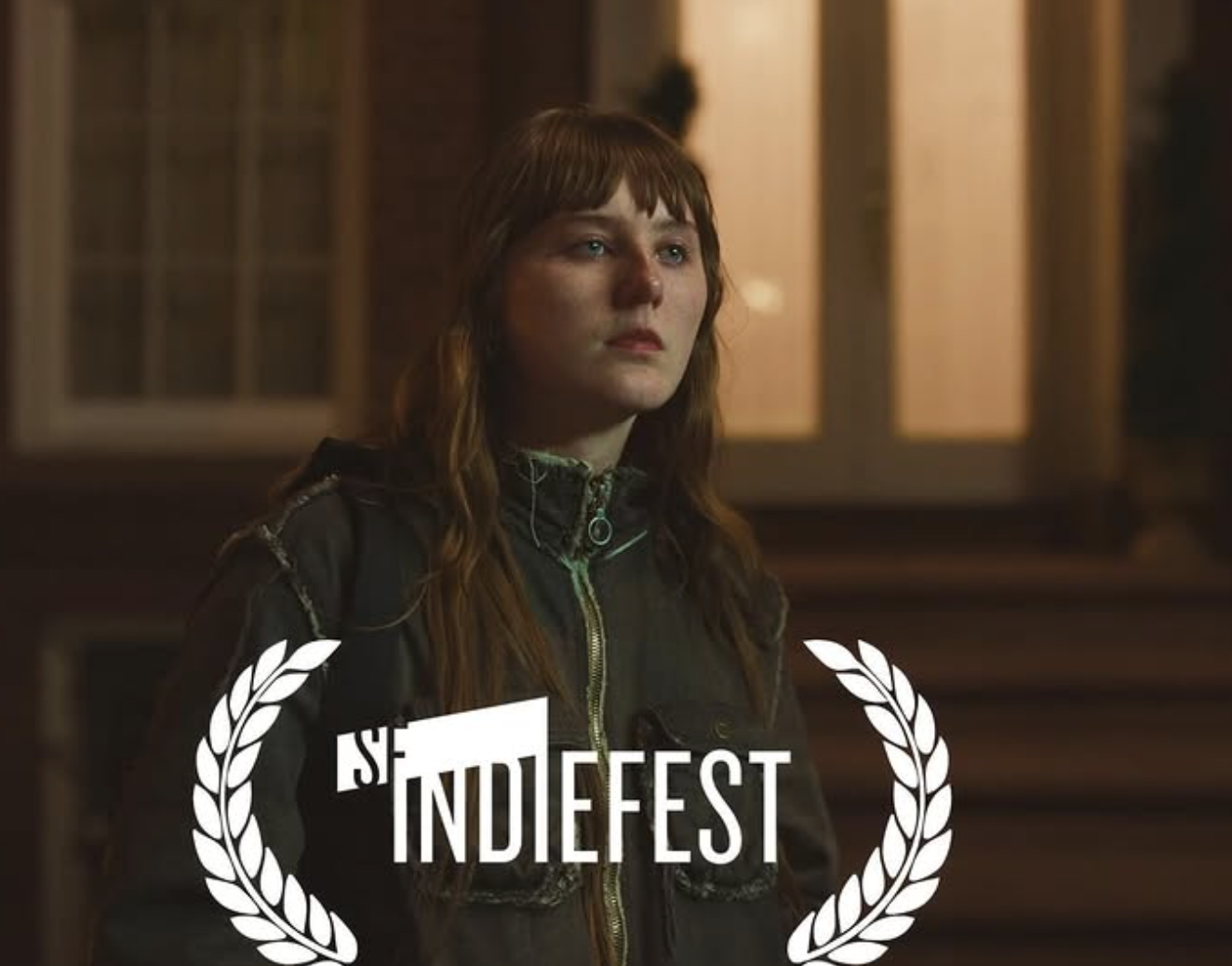 A young woman with long, wavy brown hair and blue eyes stands indoors, wearing a dark jacket with a zipper, in front of a wooden background. She has a serious or contemplative expression. The image features the text 'the INDIFFEST' surrounded by a laurel wreath.