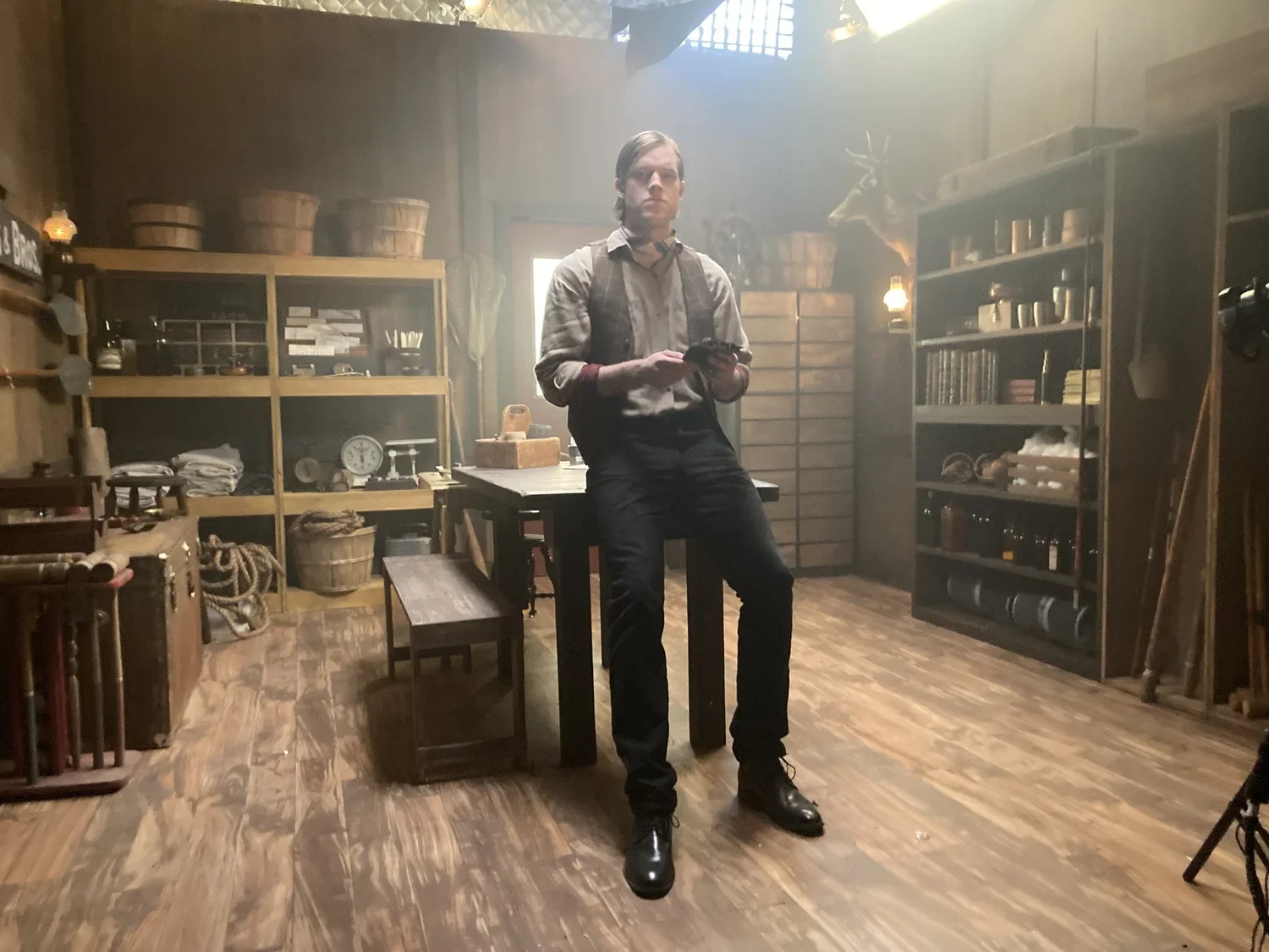 A man standing in a rustic room with wooden shelves, baskets, and vintage items, holding a phone.