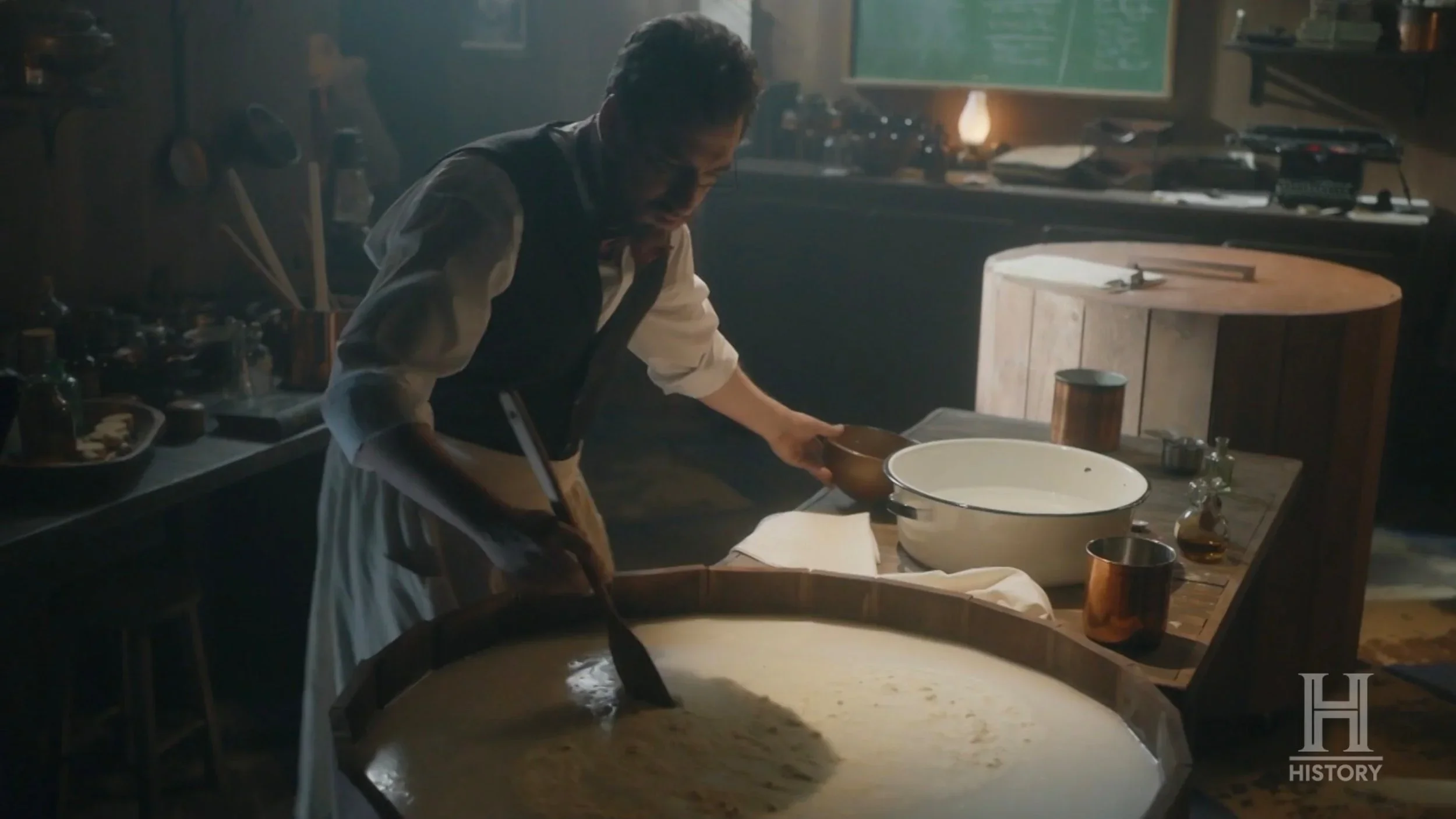 A woman cooking in a rustic kitchen with a large round wooden table and various cooking utensils and ingredients.