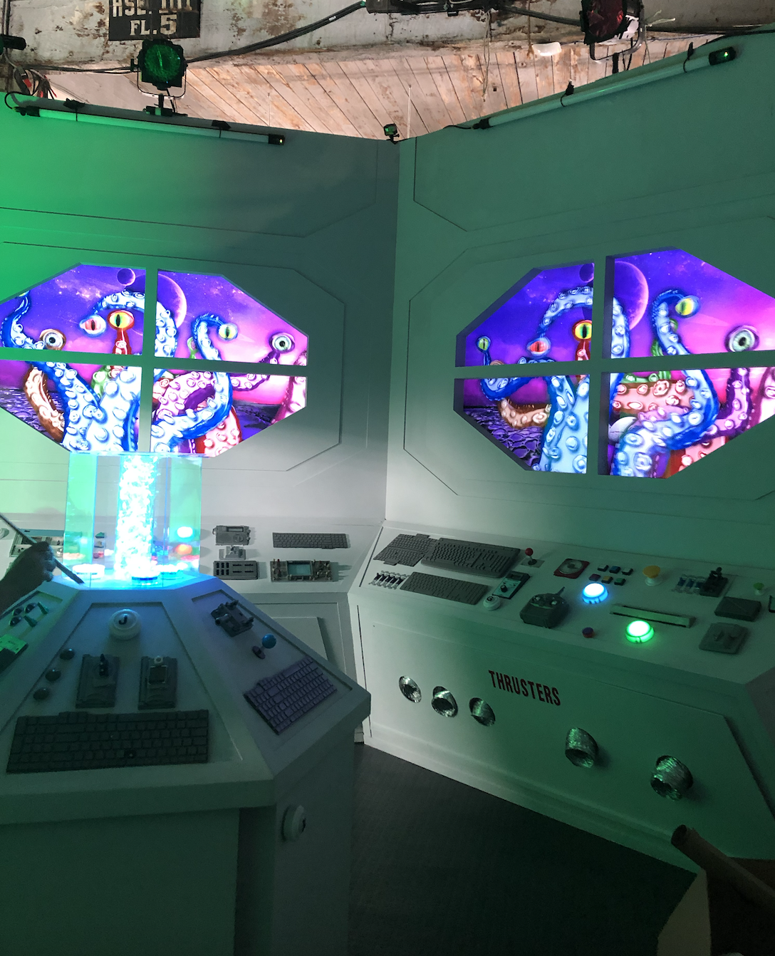 A control room with two large screens displaying animated tentacles and eyes in a colorful, sci-fi style; the room has various control panels, keyboards, buttons, and switches, with labeled thrusters.