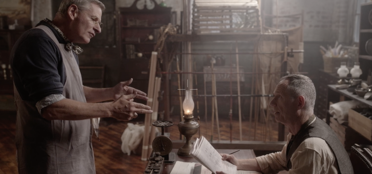 Two men engaged in a discussion in a workshop filled with tools and wooden materials. One man is standing with hands gesturing, while the other is seated, holding papers and a pencil, with a lantern and scales on the table between them.