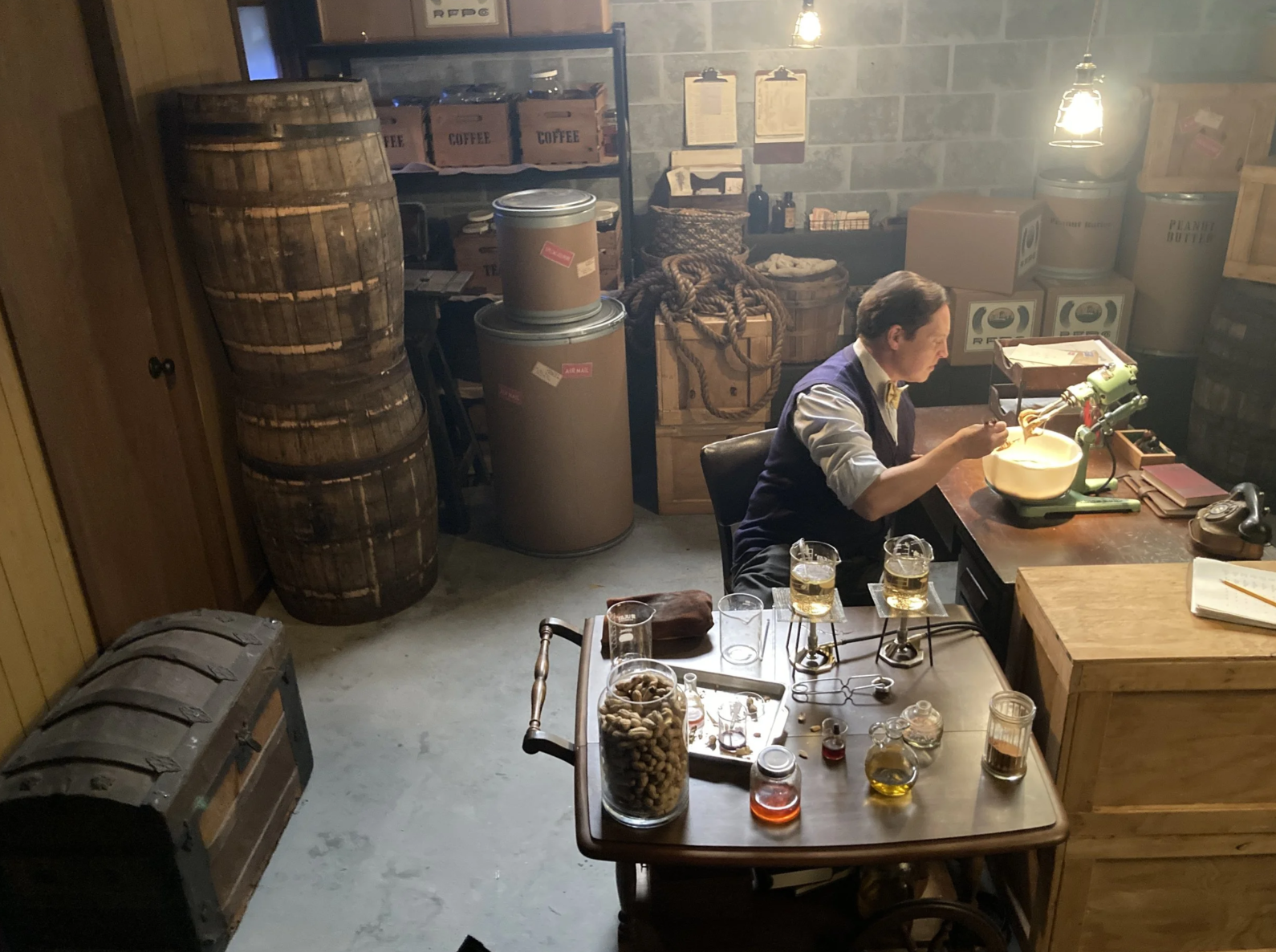 A man sitting at a wooden table working with a vintage green coffee roasting machine surrounded by jars of oils and various ingredients in a rustic café or workshop setting with barrels, boxes, and industrial lighting.