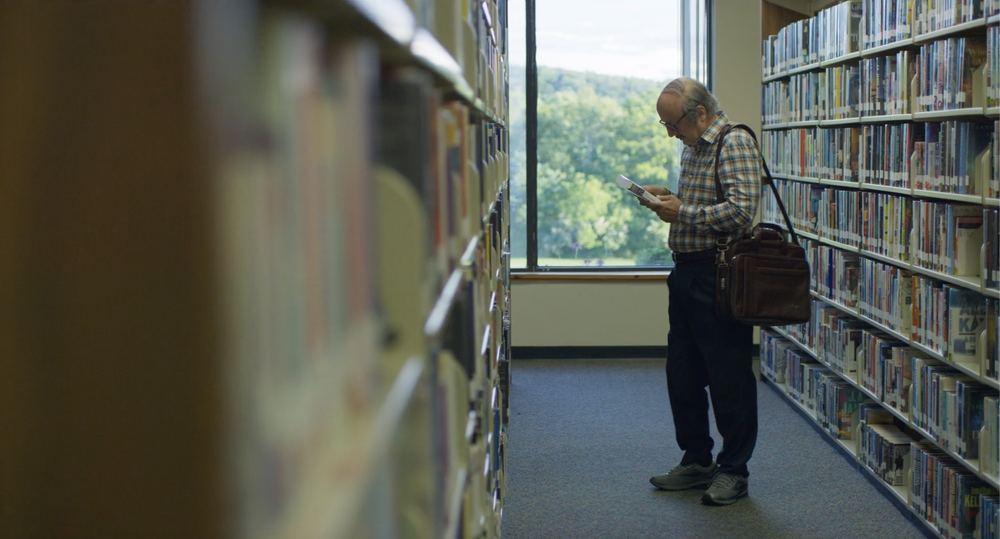 An elderly man with glasses and a checkered shirt reading a book in a library aisle, carrying a brown book bag. Large window in the background shows green trees outside.