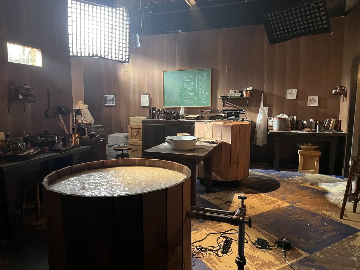 A rustic kitchen or studio space with wooden walls and a large, round wooden tub filled with water in the foreground. There are various kitchen tools, pots, and utensils on shelves and counters. A large black chalkboard with writing is mounted on the wall, and boxes and miscellaneous items are scattered around. Lighting equipment is visible overhead, suggesting it might be a set or a staged environment.
