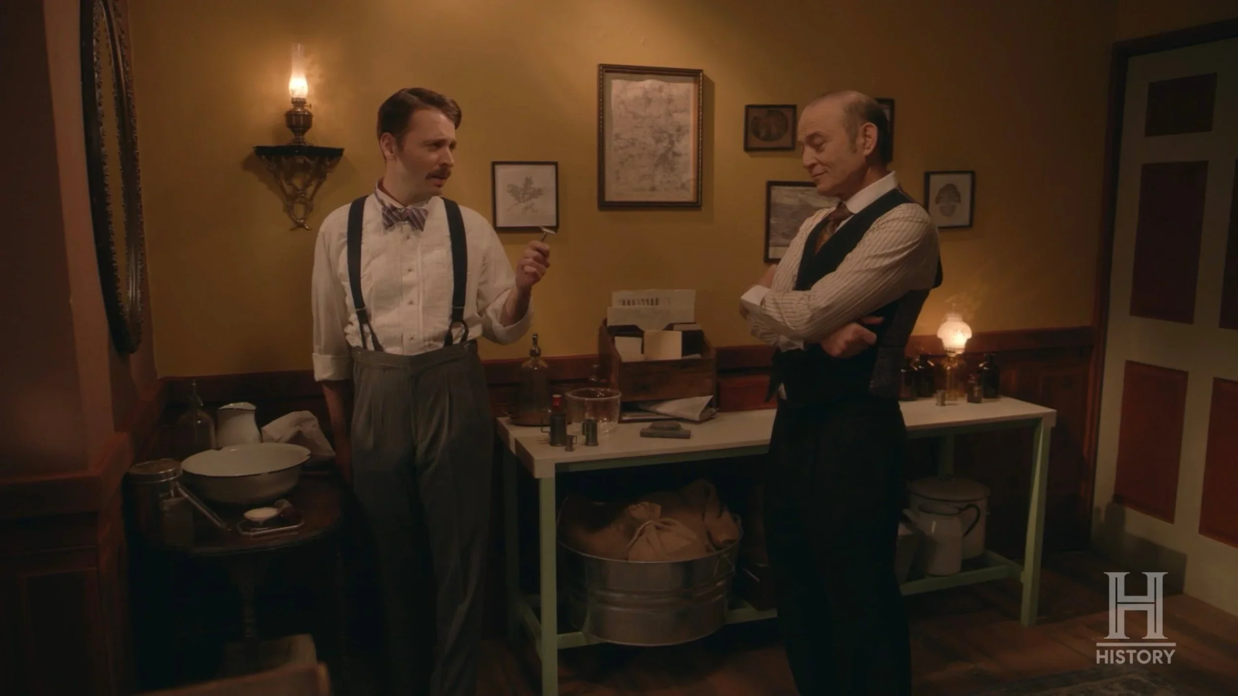 Two men having a conversation in a vintage-style room with yellow walls, framed pictures, and antique lamps. One man, with a mustache, is holding a small object and wearing suspenders and a bow tie. The other man, with his arms crossed, is dressed in a vest and striped shirt. They are standing near a table with scientific equipment and bullets. HISTORY logo in the bottom right corner.
