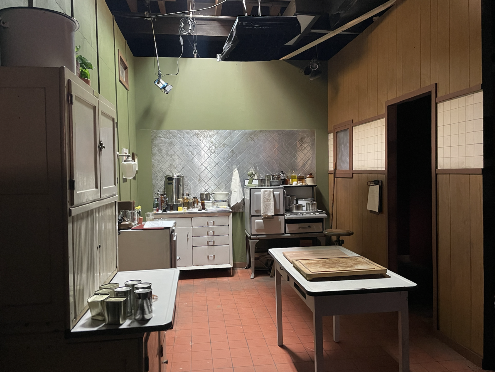 A kitchen with green and wood-paneled walls, a white counter with jars and utensils, a stove, and a table with wooden cutting boards.