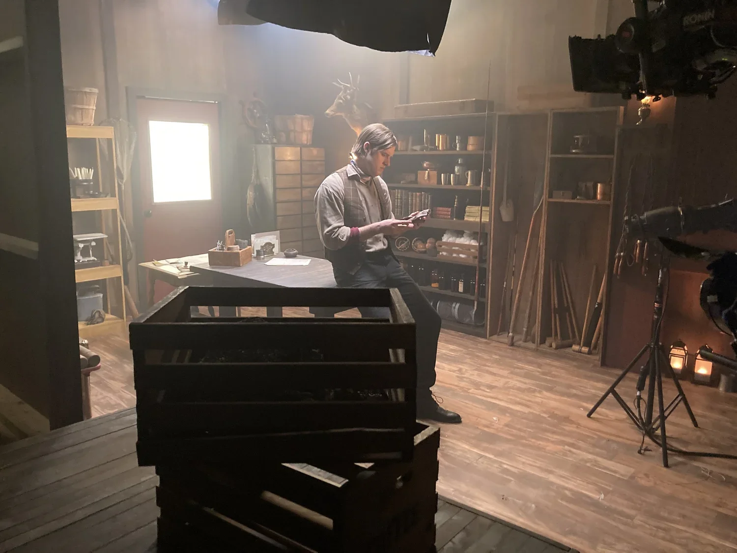 A man stands in a rustic room with wooden floors and shelves, looking at his phone, surrounded by filming equipment, candles, and wooden tools.