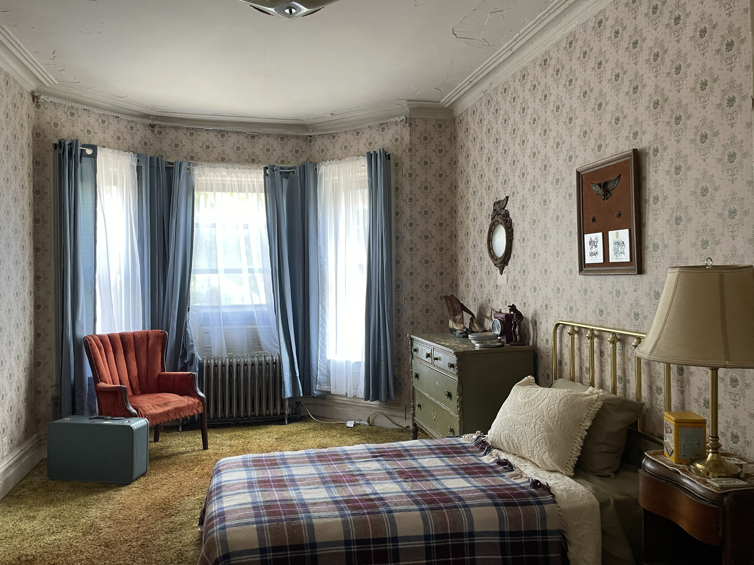 A vintage-style bedroom with floral wallpaper, a brass bed with plaid bedding, a beige pillow, a bedside table with a lamp, a green dresser with dishes and decorative items, a red velvet armchair, blue and white curtains, an orange suitcase, a radiator, and framed wall decorations.