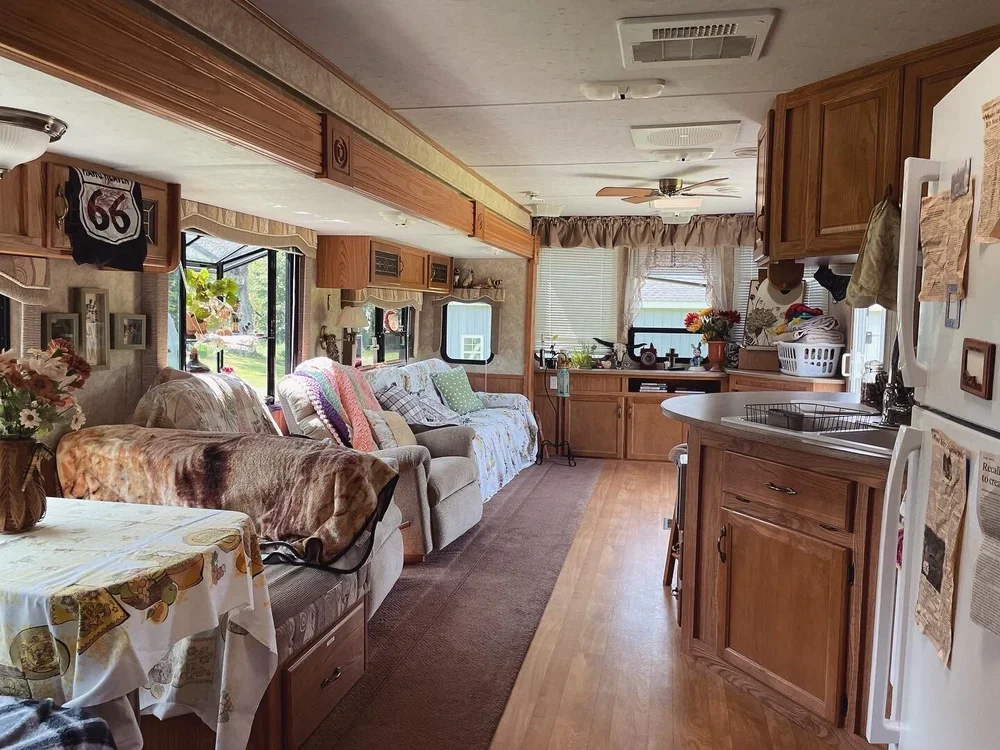 Interior of a cozy mobile home or RV, showcasing a kitchen and living area with wooden cabinets, a sofa with blankets, a table with a tablecloth, and a window with curtains.