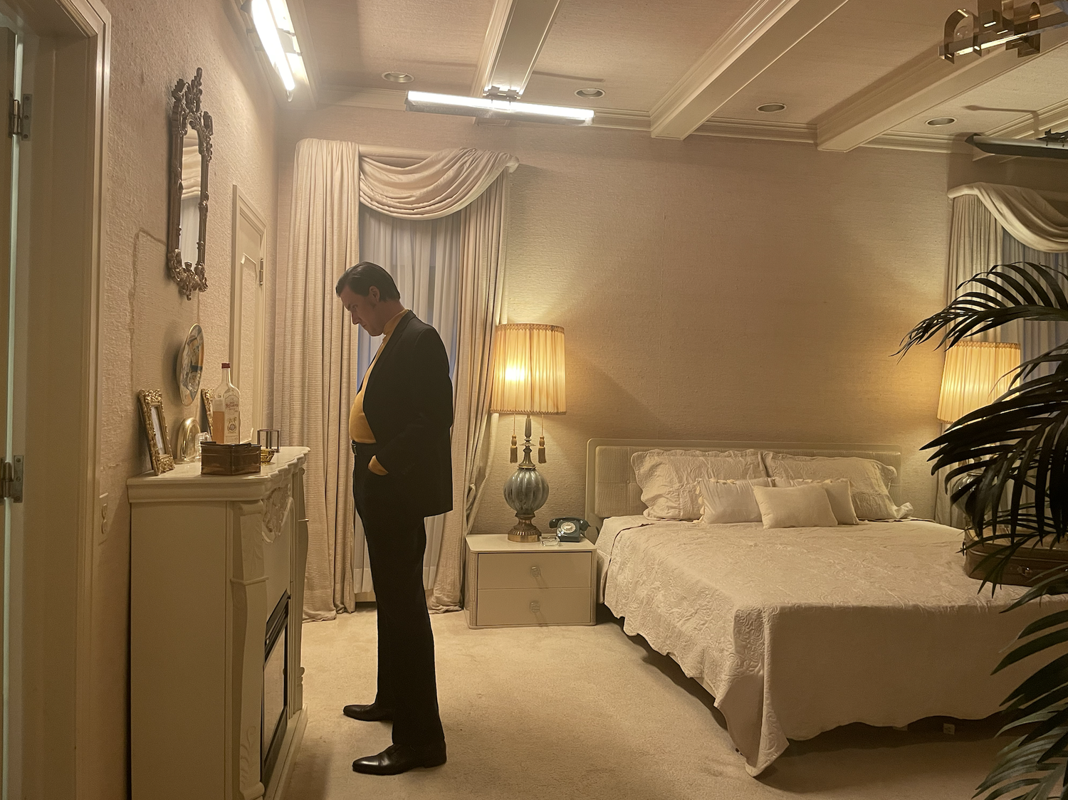 A man in a suit standing in a bedroom, looking at a dresser with framed pictures and bottles. The room has beige walls, two lamps, a bed with white bedding, and curtains.