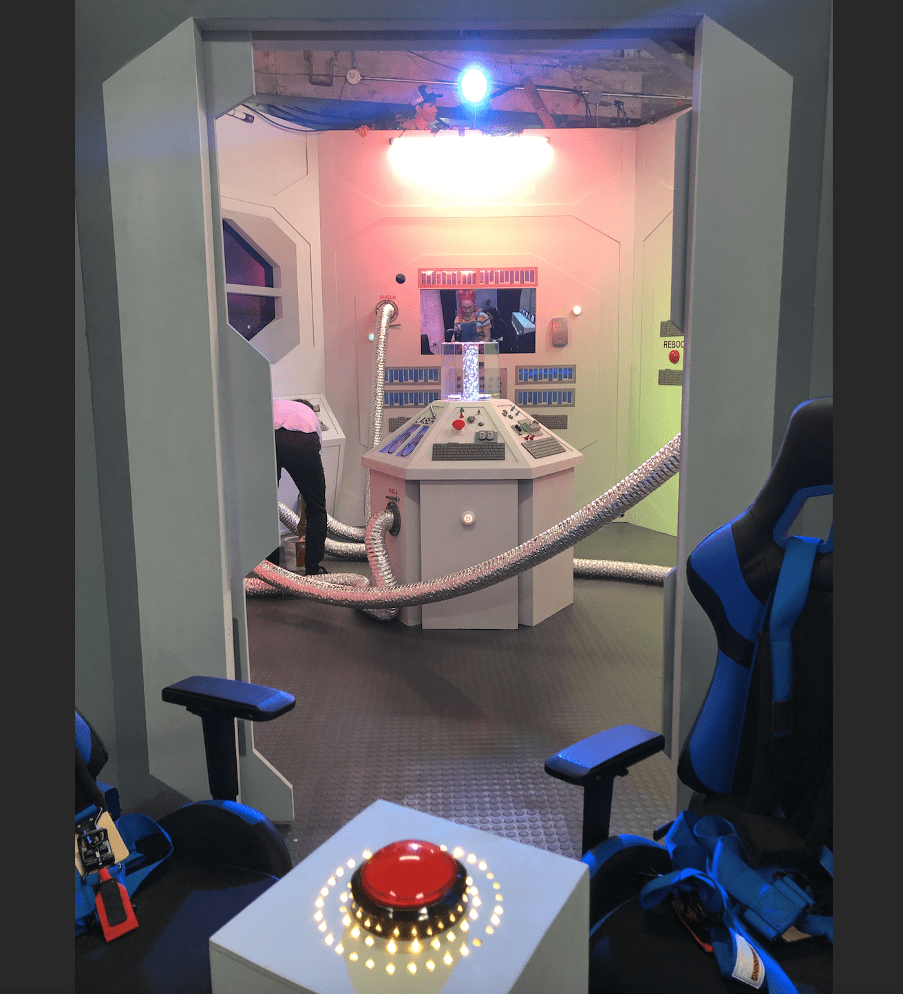 A spaceship control room with chairs, a large red button on a white pedestal, control panels, and a person with a headgear adjusting a console. The room has pipes, monitors, and colorful lighting.