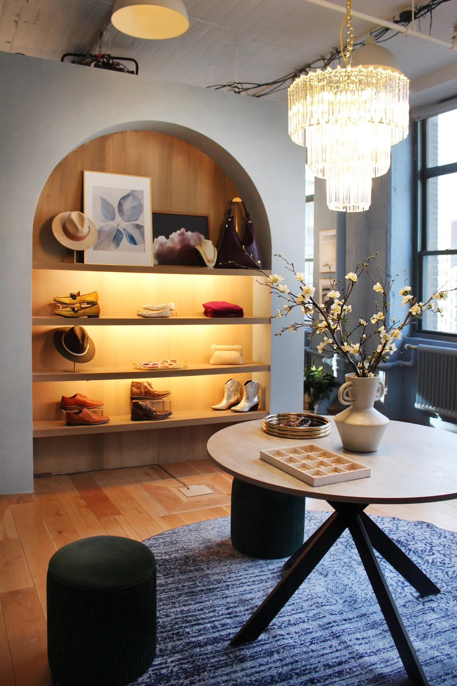 Cozy boutique with a wooden display shelf showcasing hats, shoes, and handbags, illuminated by warm lighting. A round table with a vase of flowering branches, small trays, and decorative items, placed on a woven rug near large windows and a chandelier.