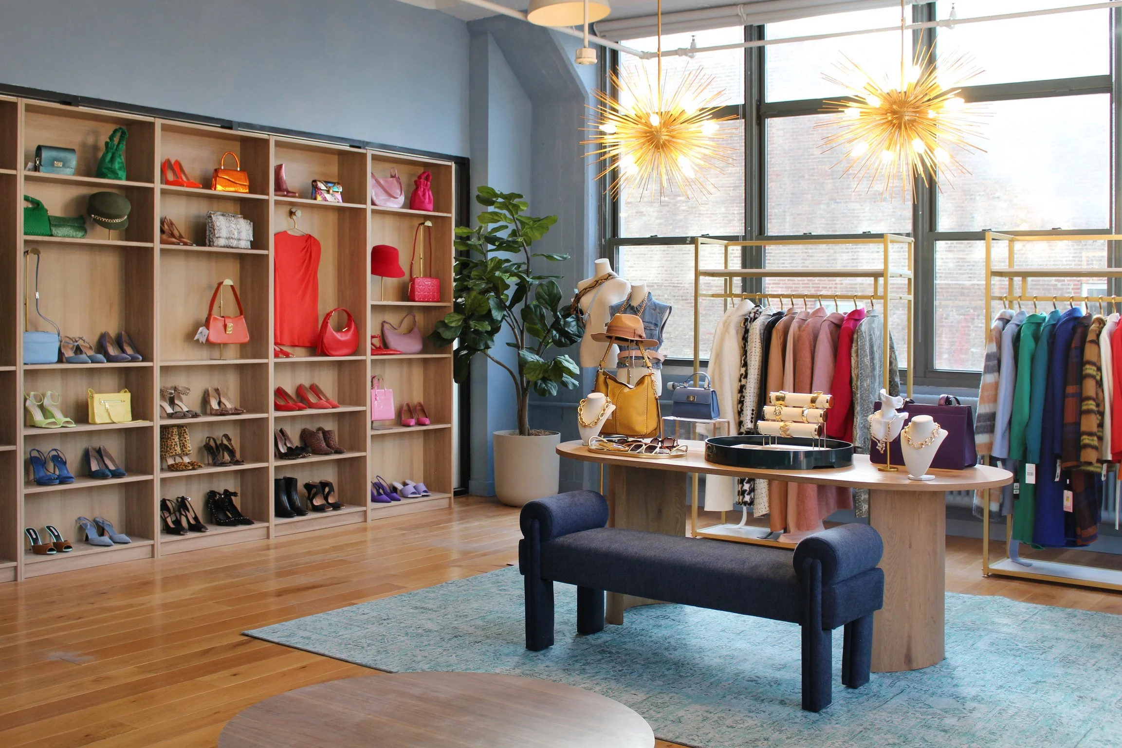 Interior of a clothing boutique with wooden shelving displaying colorful handbags and shoes, a round table with jewelry and handbags, mannequins dressed in clothing, and racks of clothes in front of large windows with natural light and modern light fixtures.