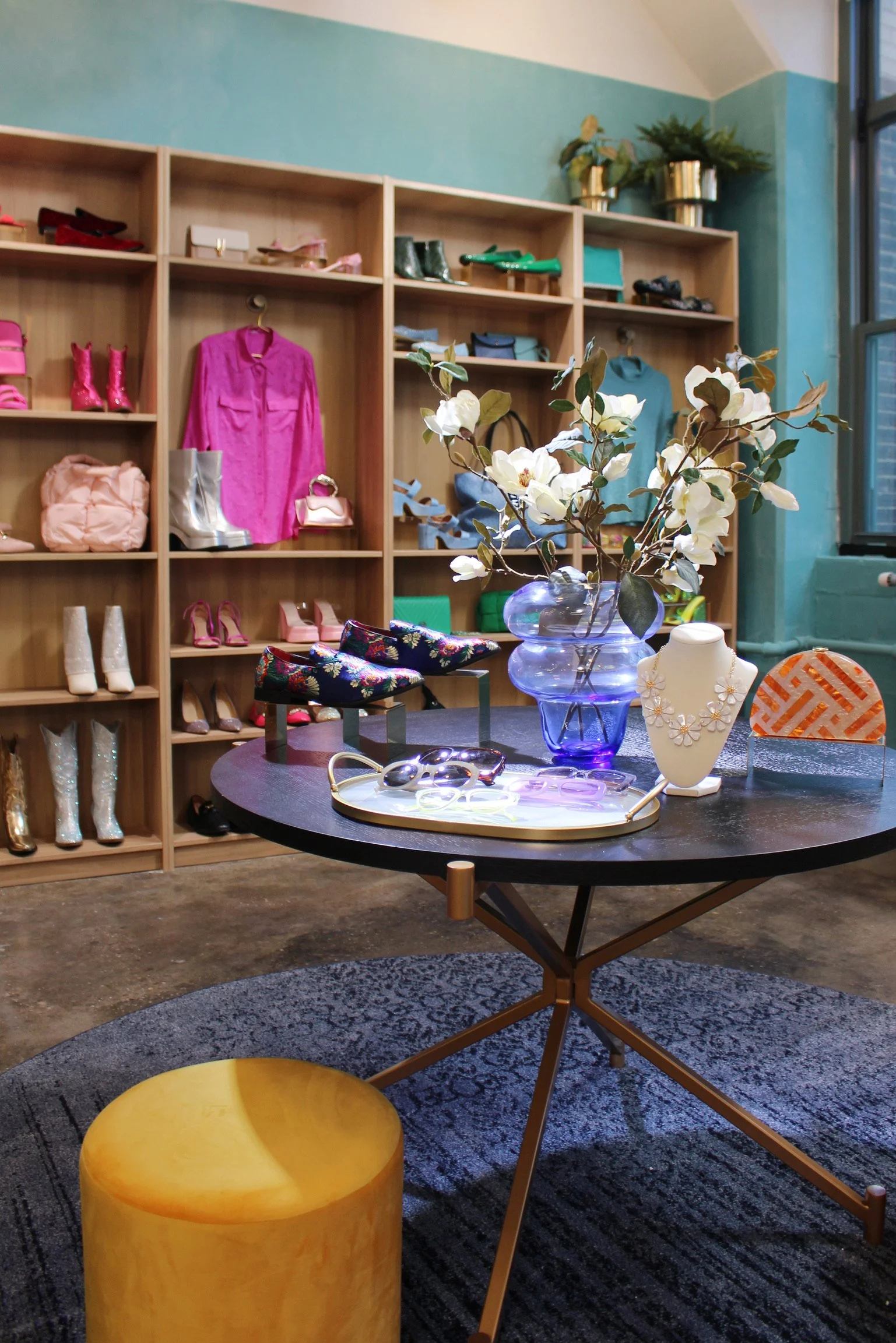 Interior view of a stylish boutique with wooden shelves displaying colorful shoes, handbags, and clothing. A round table in the foreground has a large blue vase with white flowers, a jewelry display, and decorative items. A yellow ottoman is partially visible at the base of the table. The room has a concrete floor, patterned rug, and a large window with natural light.