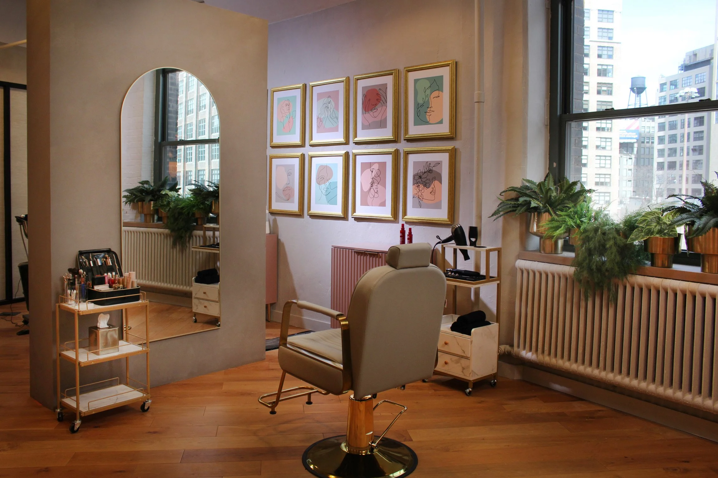 A makeup station in a salon with a large mirror, a beige salon chair, a small rolling cart with makeup supplies, framed abstract art on the wall, and large windows with city buildings outside, decorated with potted plants.